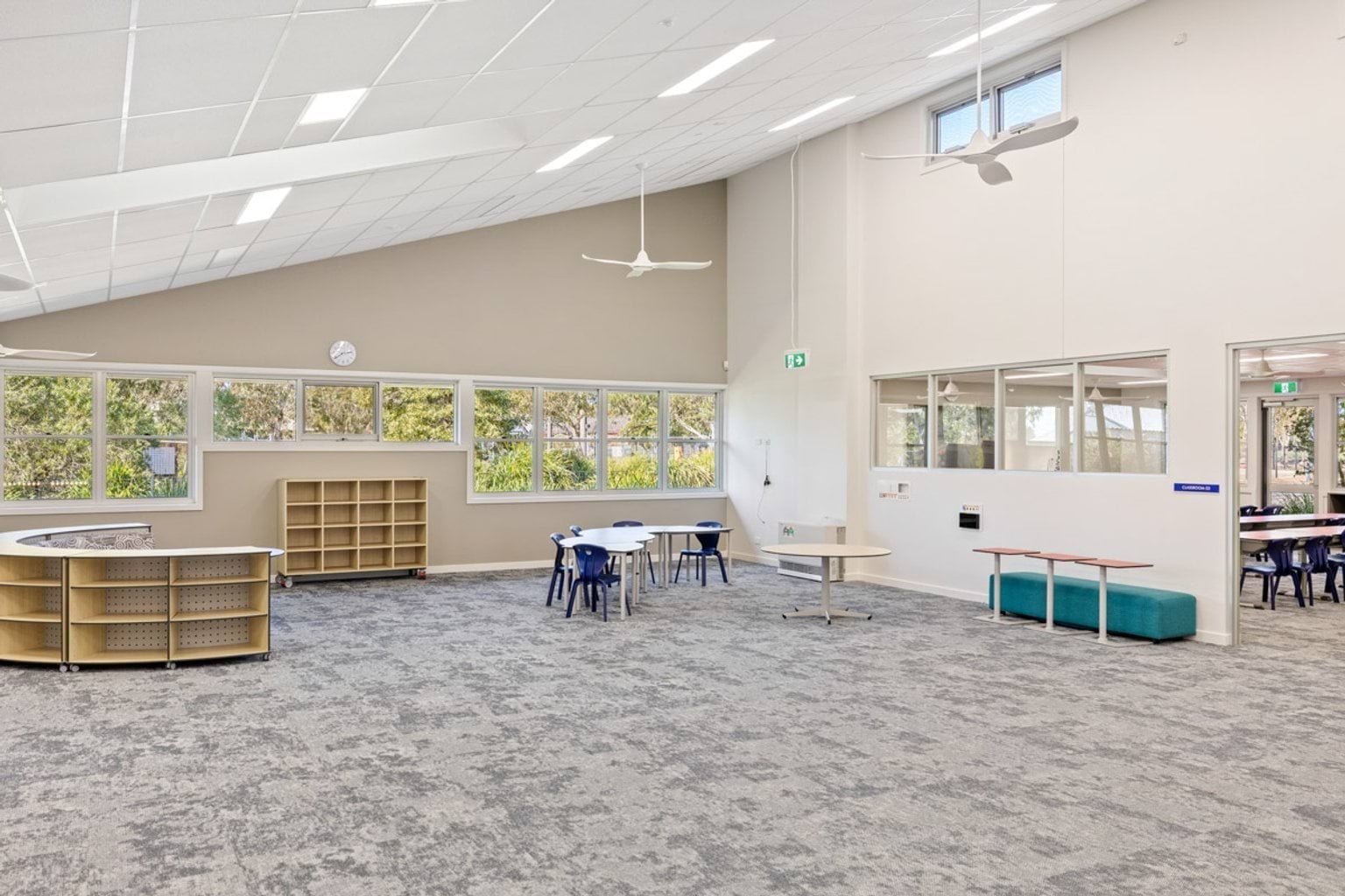 A spacious classroom with grey carpet, white walls, and modular shelving units. Large windows provide natural light.
