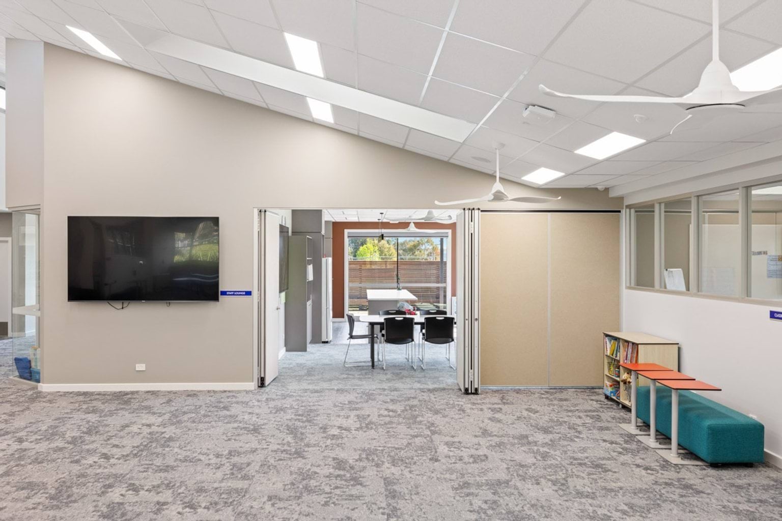 An open learning space with a wall-mounted TV, grey carpet, and a view into a meeting room with tables and chairs.