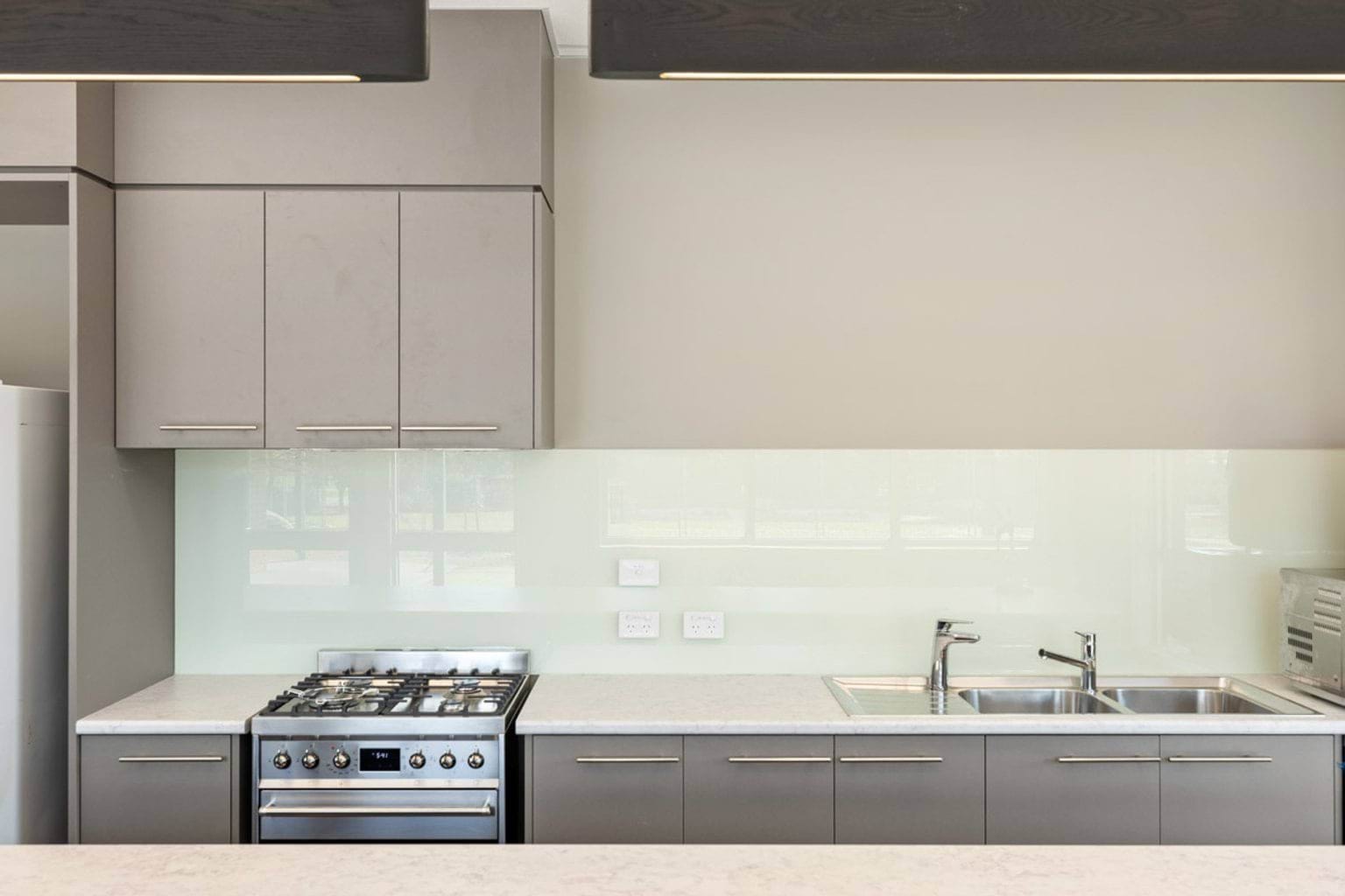 A modern kitchen with grey cabinetry, a stainless steel gas stove, double sink, and a pale glass splashback.