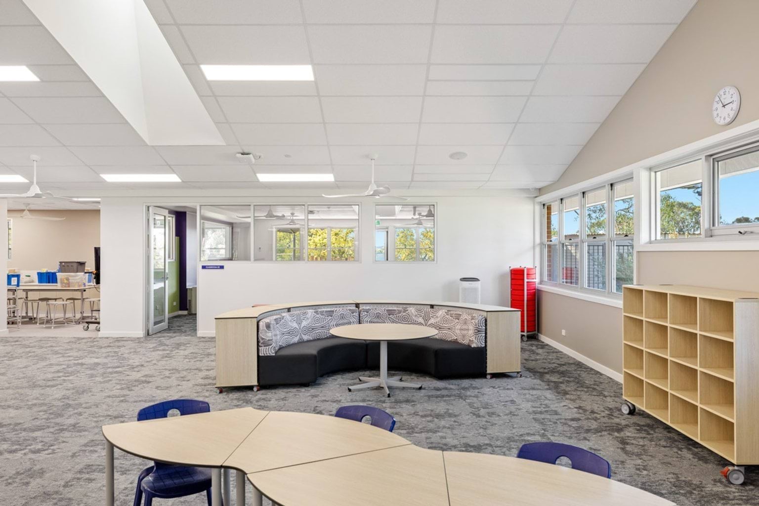 An open learning area with curved upholstered seating, tables, and shelving units. Windows overlook greenery.