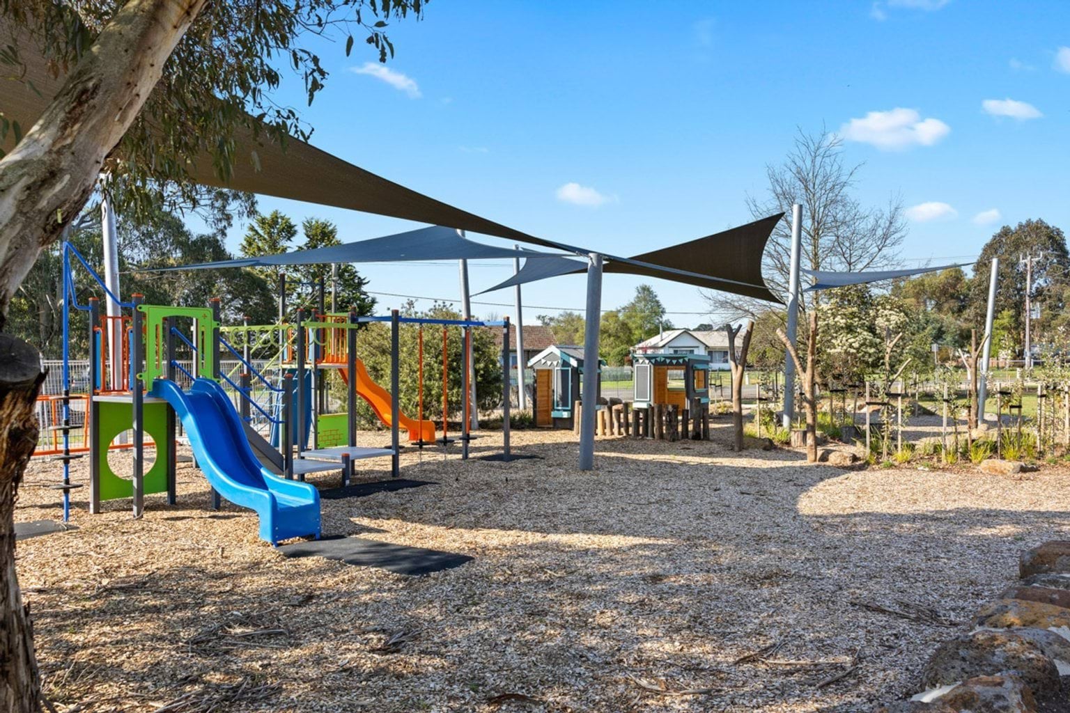A playground featuring bright slides, climbing structures, and shade sails, surrounded by landscaped garden beds.