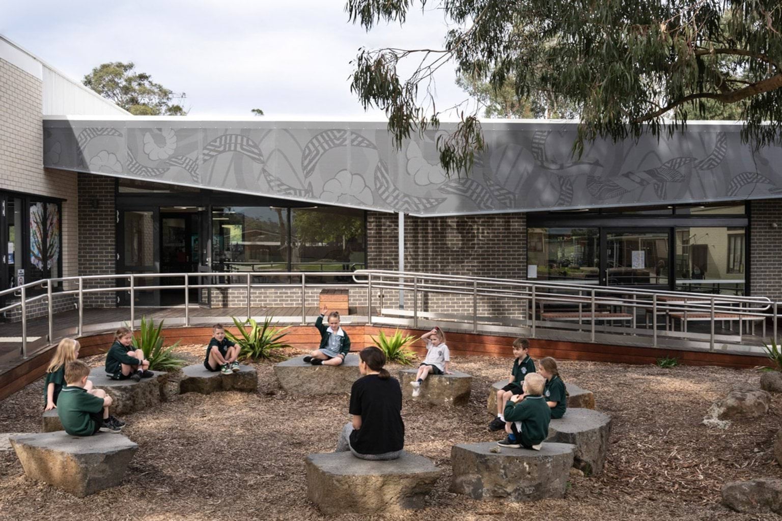 Students sitting on large rocks in an outdoor learning space with timber decking and garden beds. The building has dark brick walls and perforated metal screens.