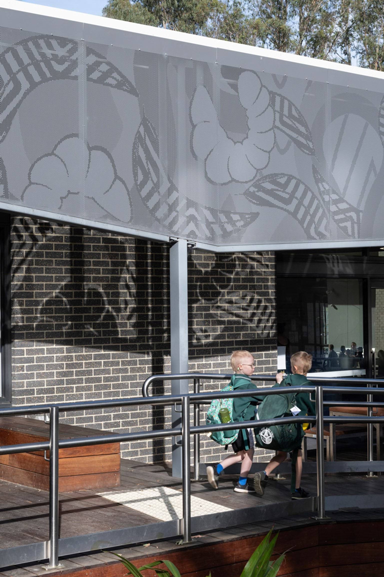 Two students walking up a timber ramp with stainless steel handrails under a perforated metal shade panel featuring decorative patterns.