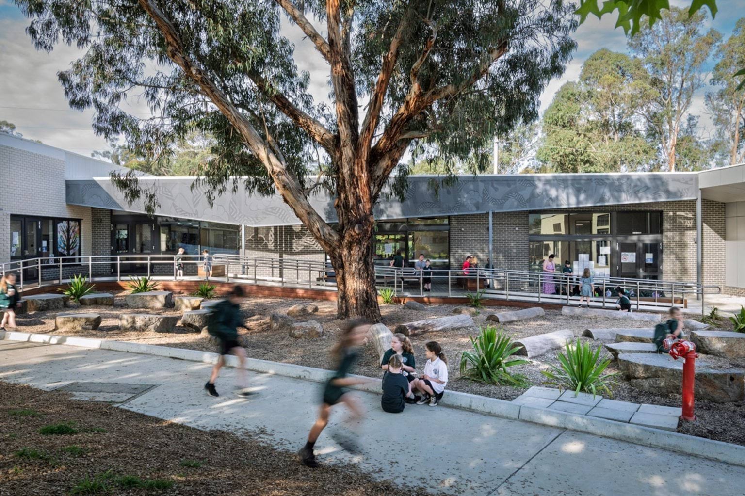 Students running and sitting in a landscaped outdoor area with a large gum tree, timber decking and garden beds. The building has dark brick walls and perforated screens.