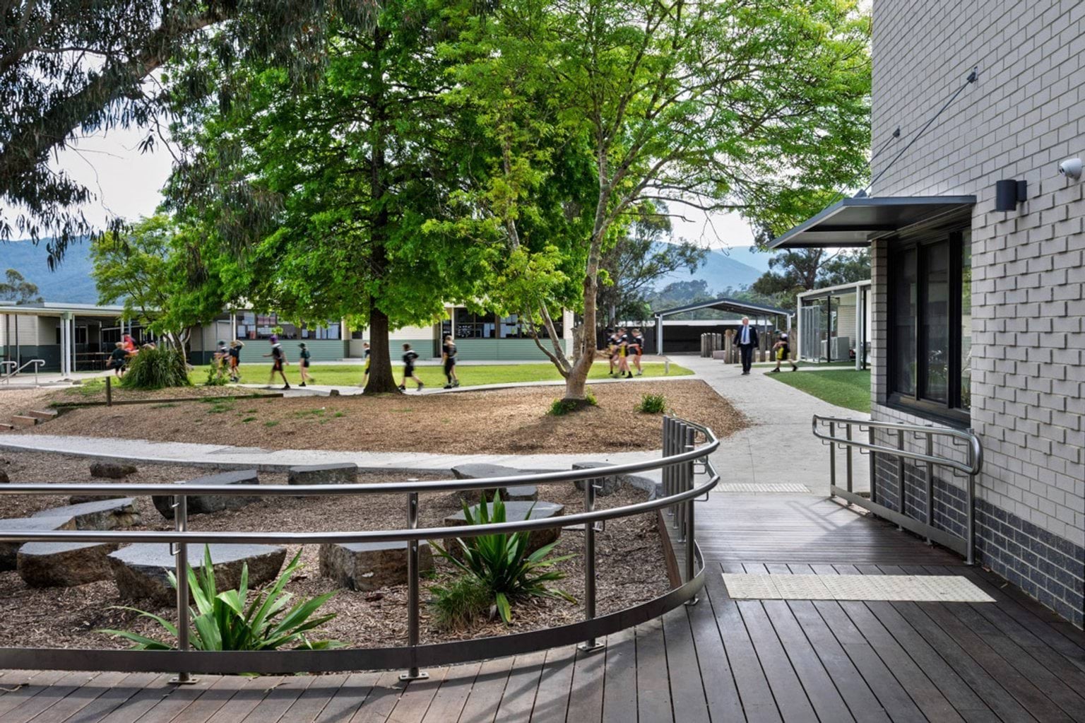 A landscaped outdoor area with timber decking, curved metal handrails and garden beds. Large trees provide shade and classrooms are visible in the background.