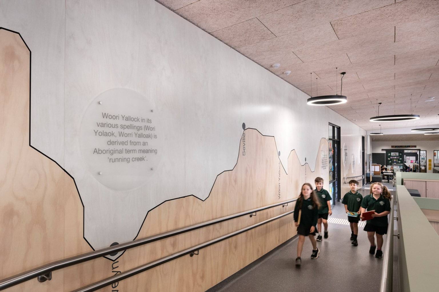 Students walking along an indoor ramp beside a wall mural with local place names and Aboriginal language details. Circular pendant lights hang from the ceiling.