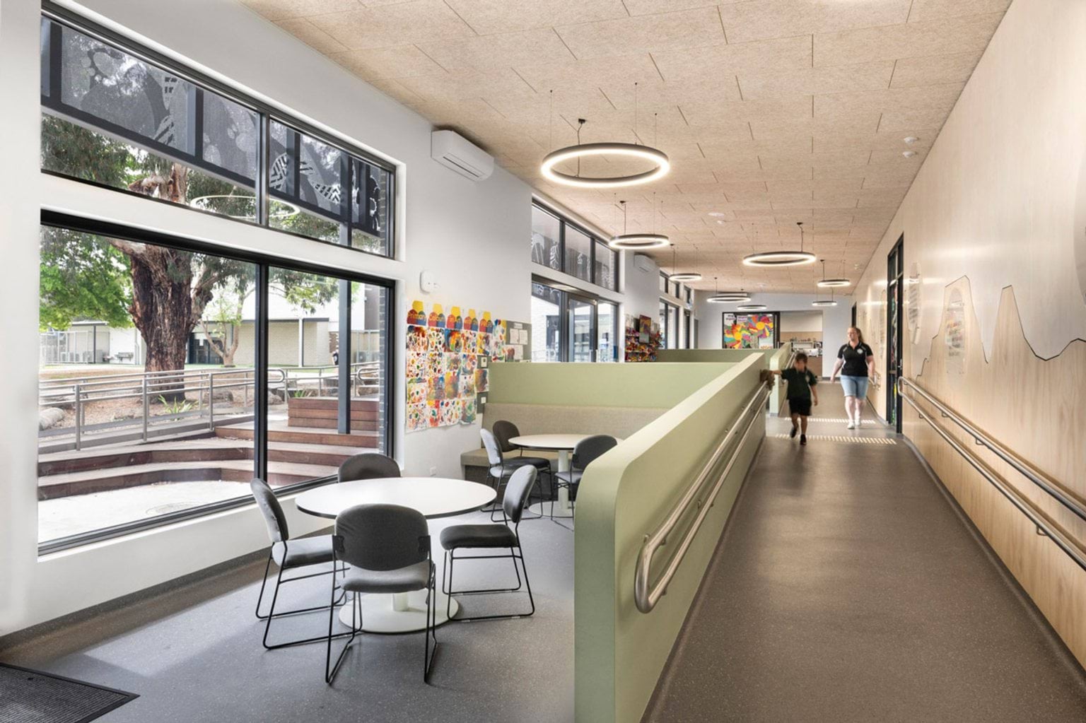 A bright indoor space with round tables and chairs near large windows overlooking a landscaped courtyard. A ramp runs along the wall with a mural of local landmarks.