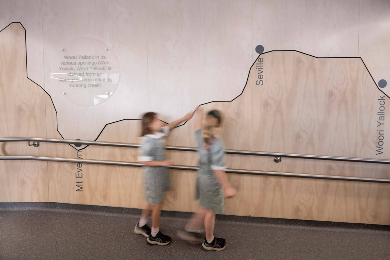 Two students walking along an indoor ramp beside a wall mural showing a map of local towns and mountains, with Aboriginal language details.