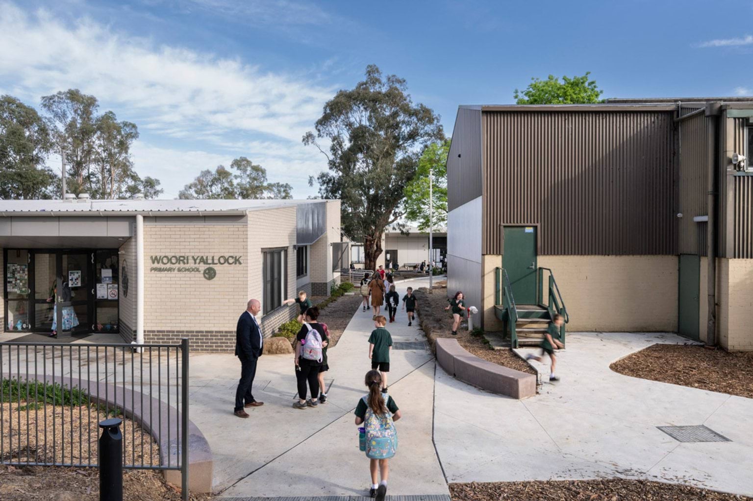 The main entrance of Woori Yallock Primary School with cream brick walls and signage. Students and staff are walking along concrete paths between buildings.