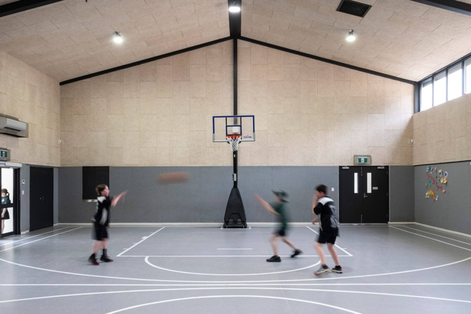 Students playing basketball inside a school gymnasium with a high pitched ceiling, light timber wall panels and a grey sports floor marked with court lines.