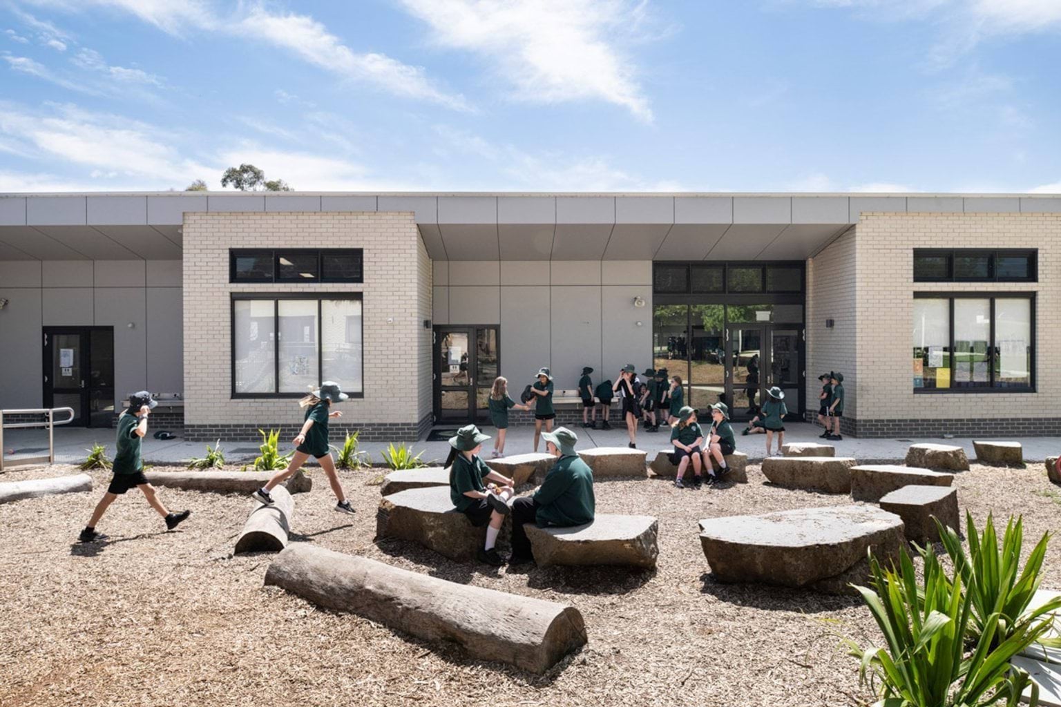Students playing and sitting on large rocks in a courtyard with timber logs and garden beds. The building behind has cream brick walls and large windows.