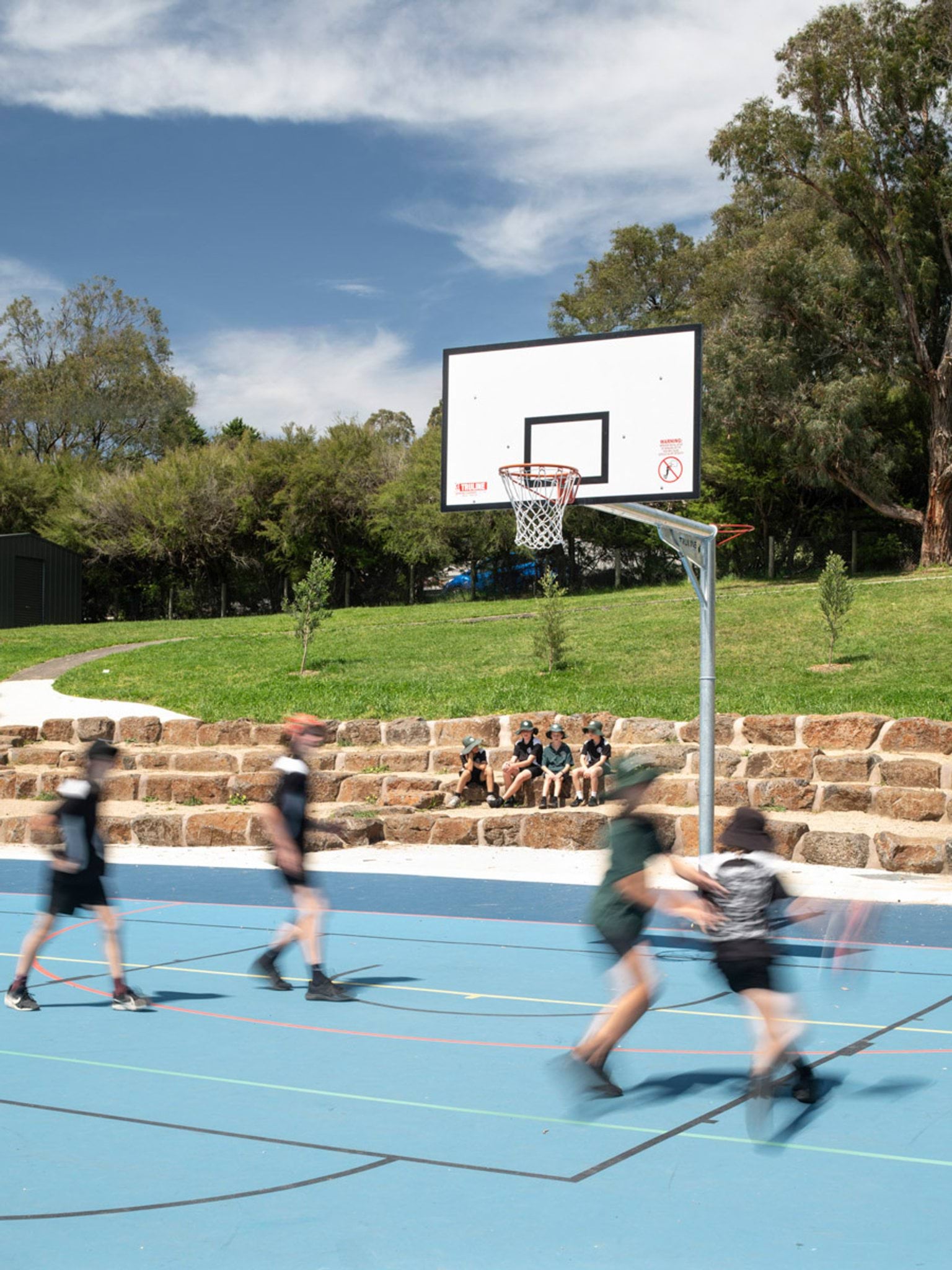 Students playing basketball on an outdoor blue sports court with a stone retaining wall and tiered seating. Trees and a grassy hill surround the area.