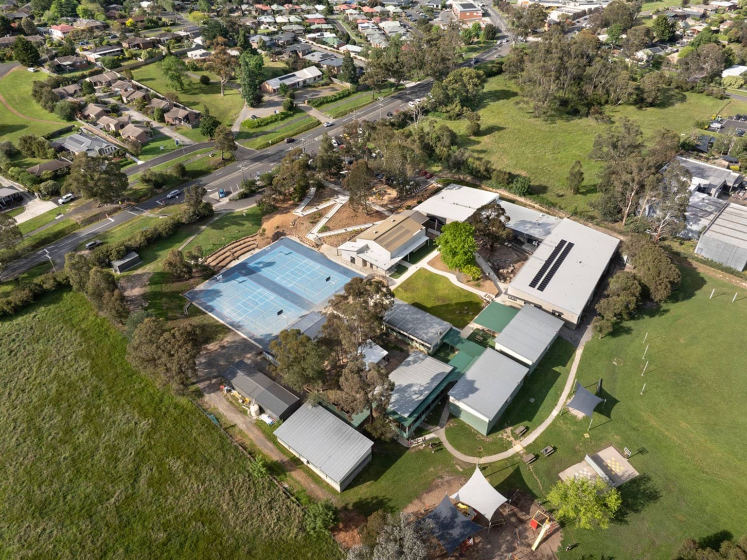 An aerial view of Woori Yallock Primary School showing multiple buildings, a large blue sports court, landscaped outdoor areas and surrounding residential streets.