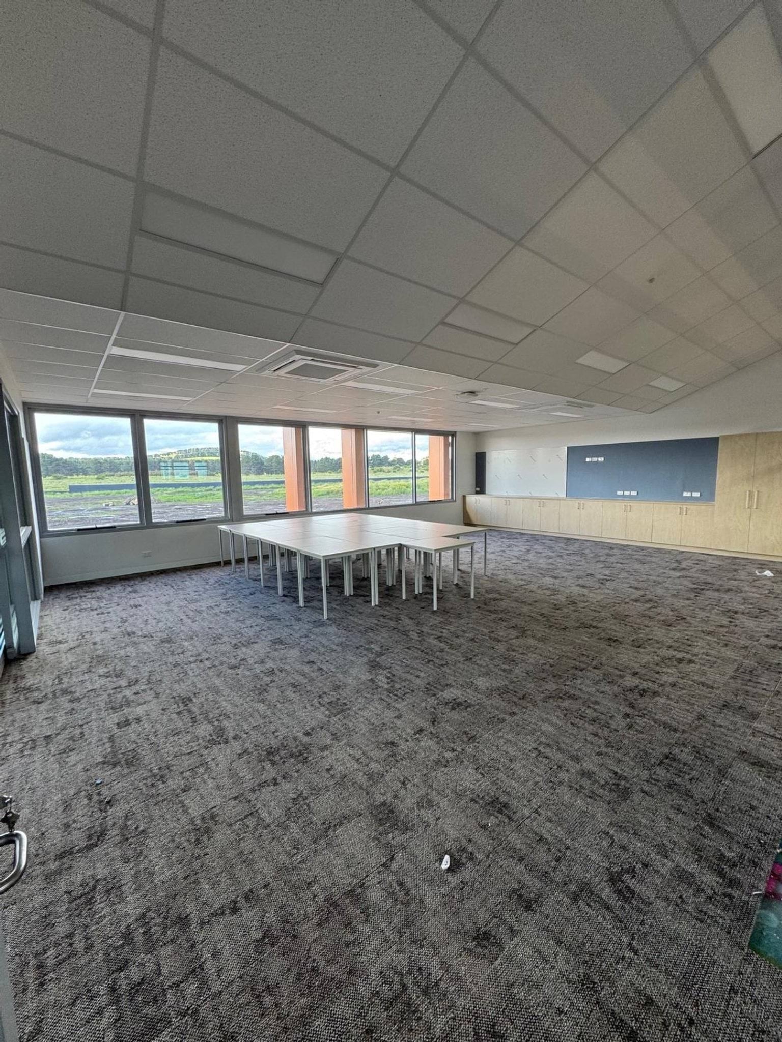 Tables in classroom next to window. There are boards and cupboards at the front of the room.