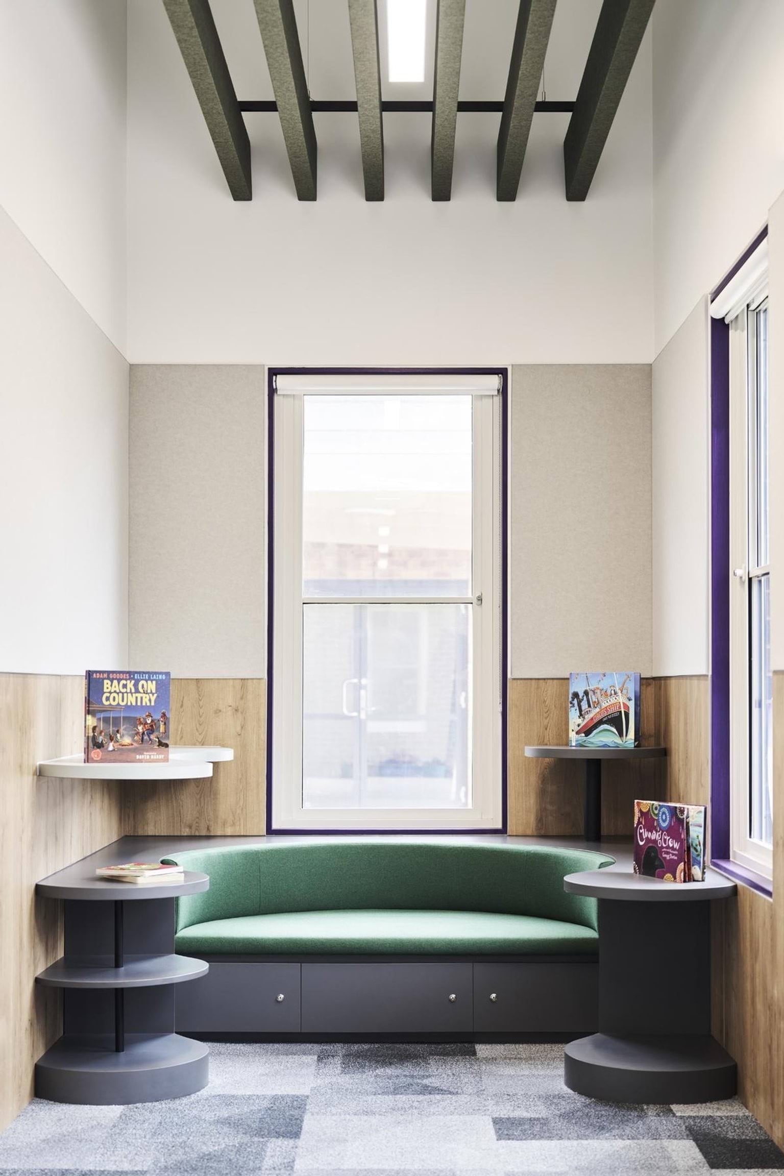 A reading nook with a curved green bench, round shelving units, and books displayed on wall-mounted shelves beneath a large window.