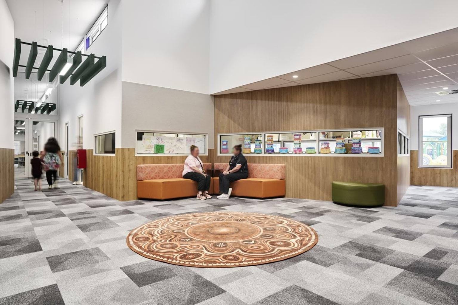 An interior space with patterned grey carpet, timber wall panels, and orange cushioned seating. Display windows with books and artwork line the wall.