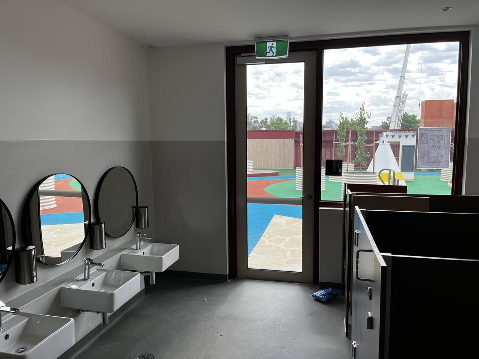 A kindergarten bathroom with three small sinks and round mirrors, next to a door leading to an outdoor play area with colourful flooring.