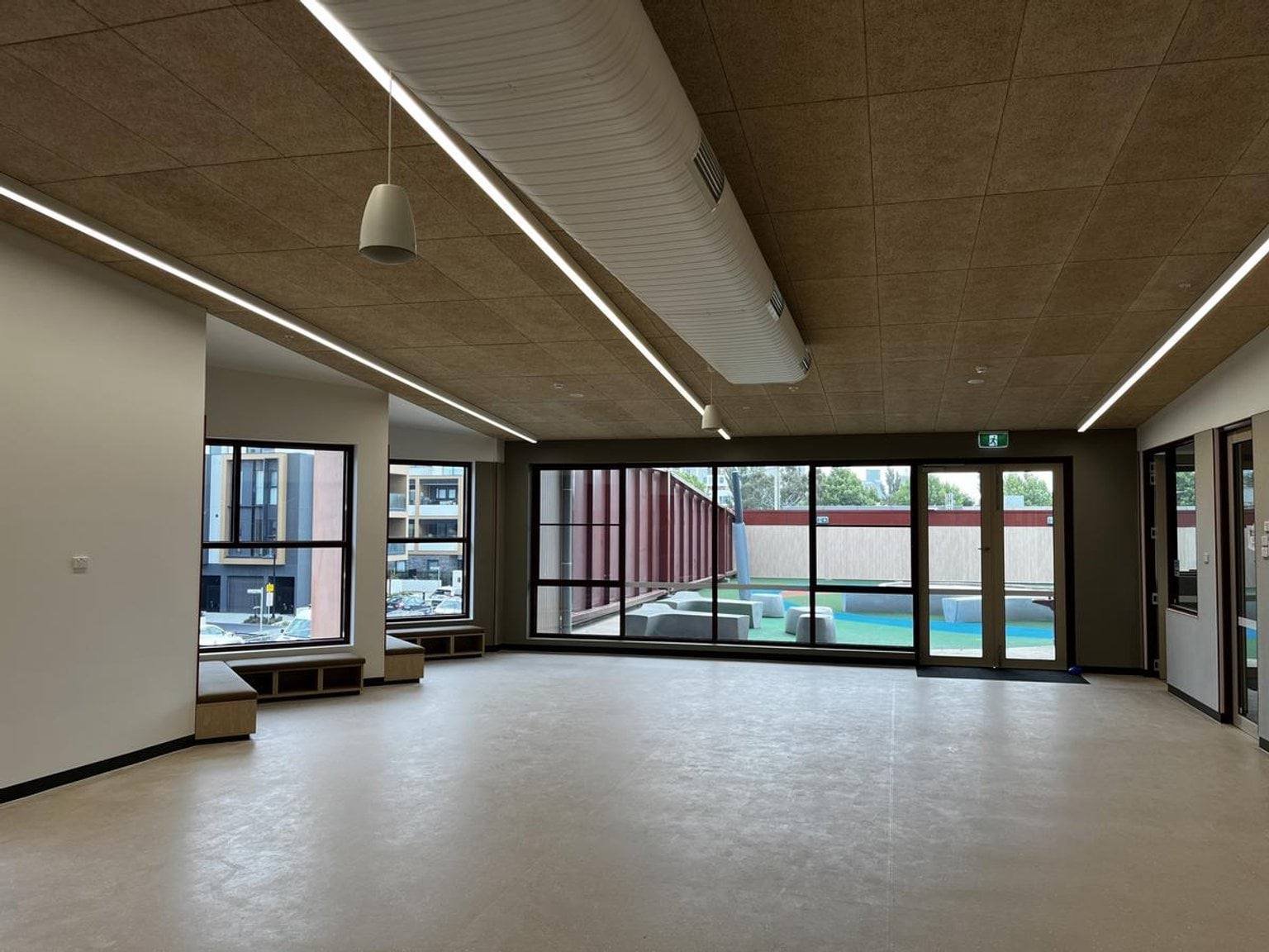 An empty kindergarten room with light-coloured flooring, timber ceiling panels, and large windows opening to an outdoor play area.