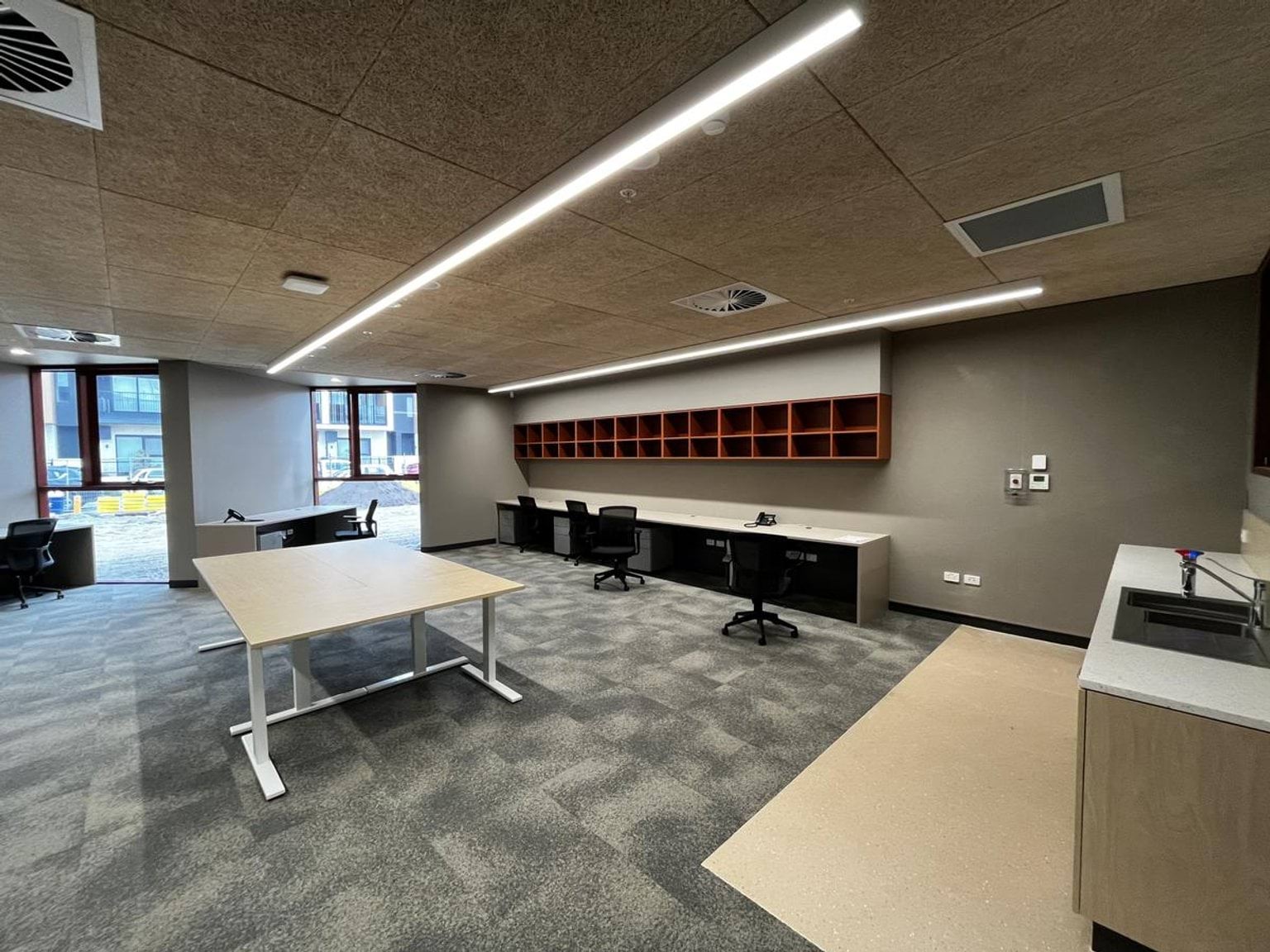 An unfinished interior room with workstations, tables, and shelving units. Large windows overlook the construction site outside.