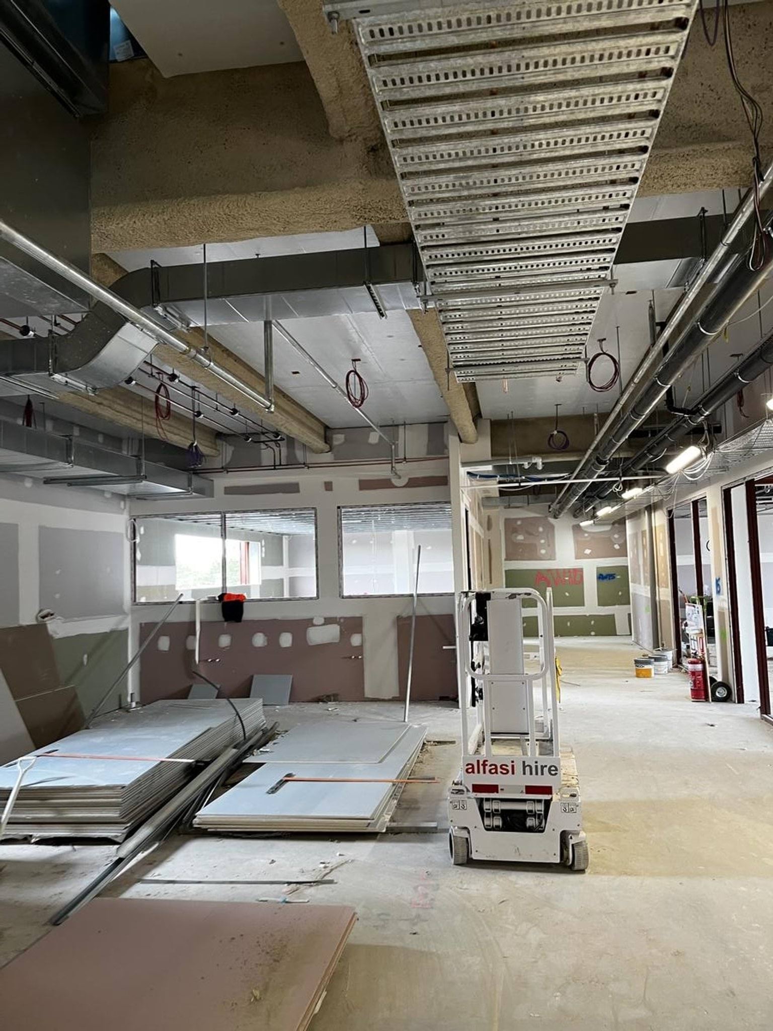 An unfinished interior space with exposed ceiling ducts, wiring, and wall panels. Construction materials and equipment are on the floor.