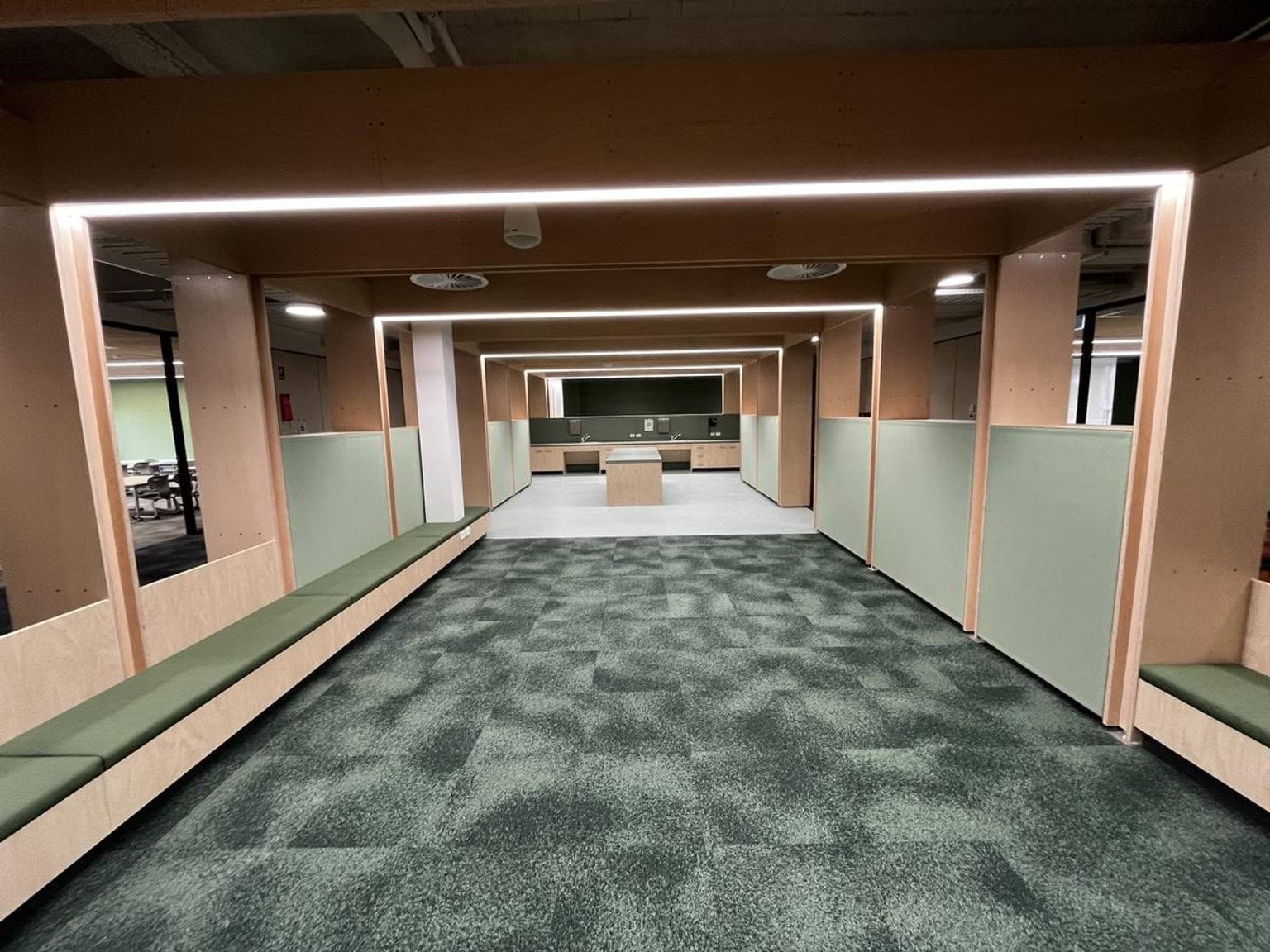 A wide interior space with green carpet, timber seating along the walls, and light panels framing the ceiling.