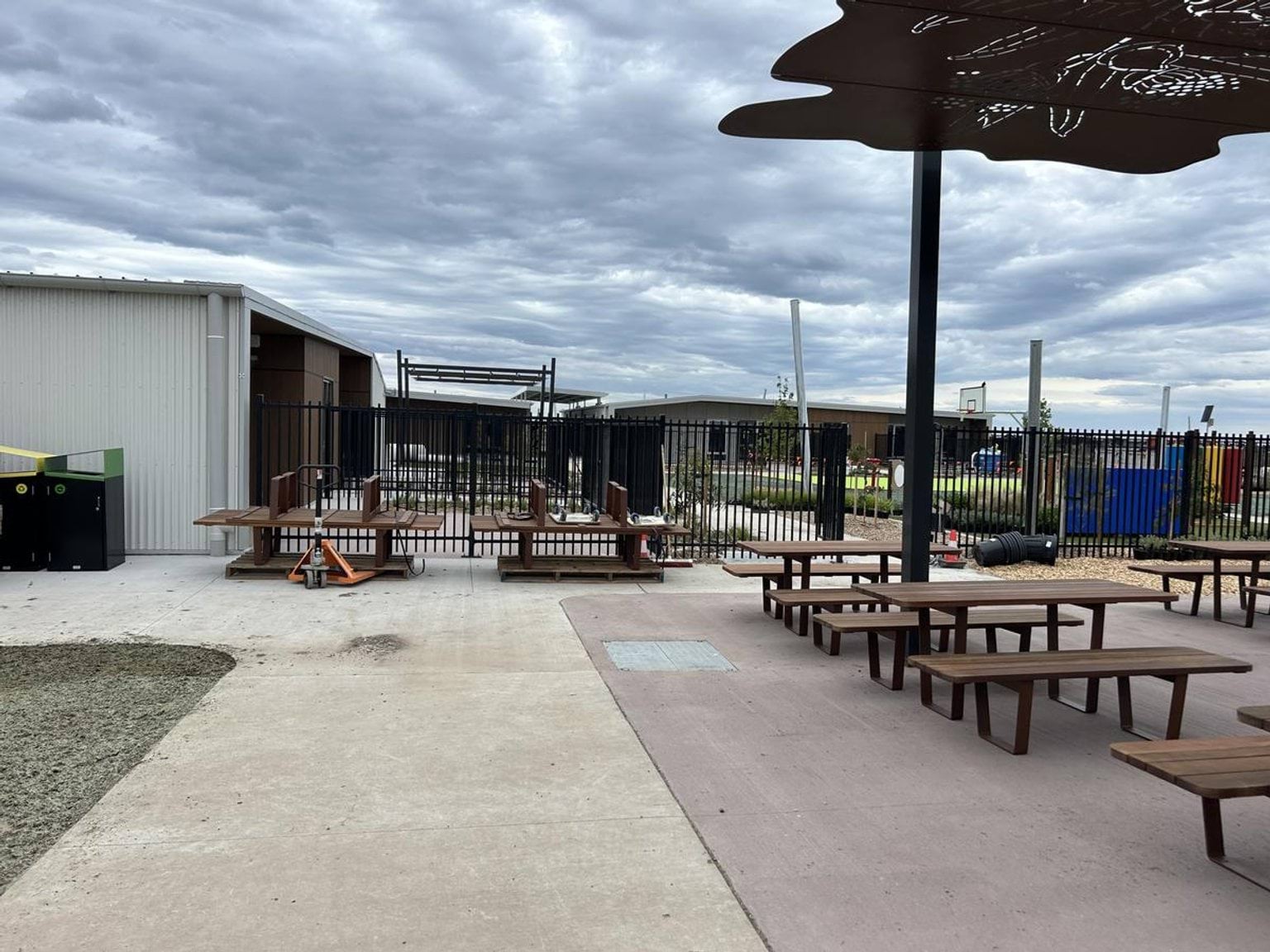 An outdoor area with picnic tables on artificial grass, a concrete path, and a playground with climbing equipment in the background.