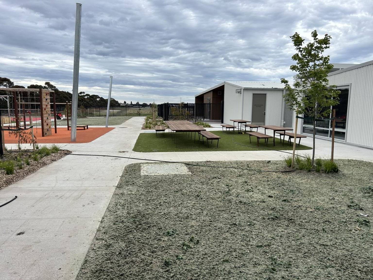 An outdoor area with picnic tables on artificial grass, a concrete path, and a playground with climbing equipment in the background.