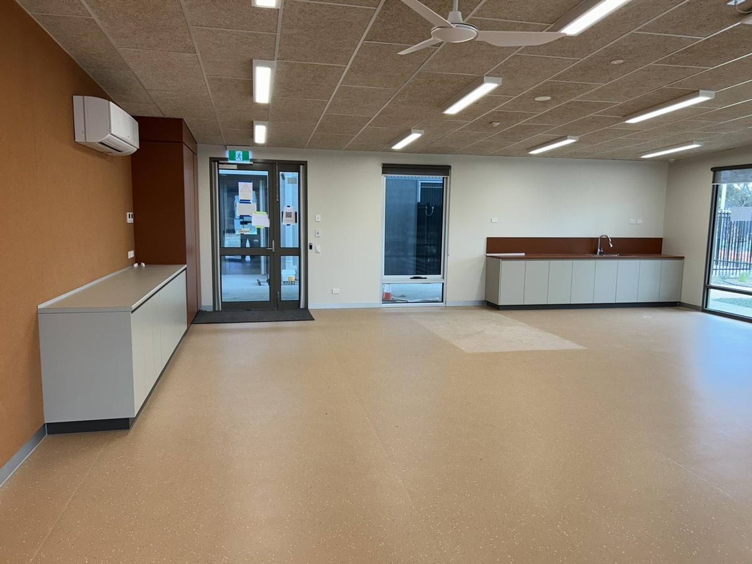 A large classroom with light flooring, built-in cabinetry, and multiple doors leading outside, under bright ceiling lights.