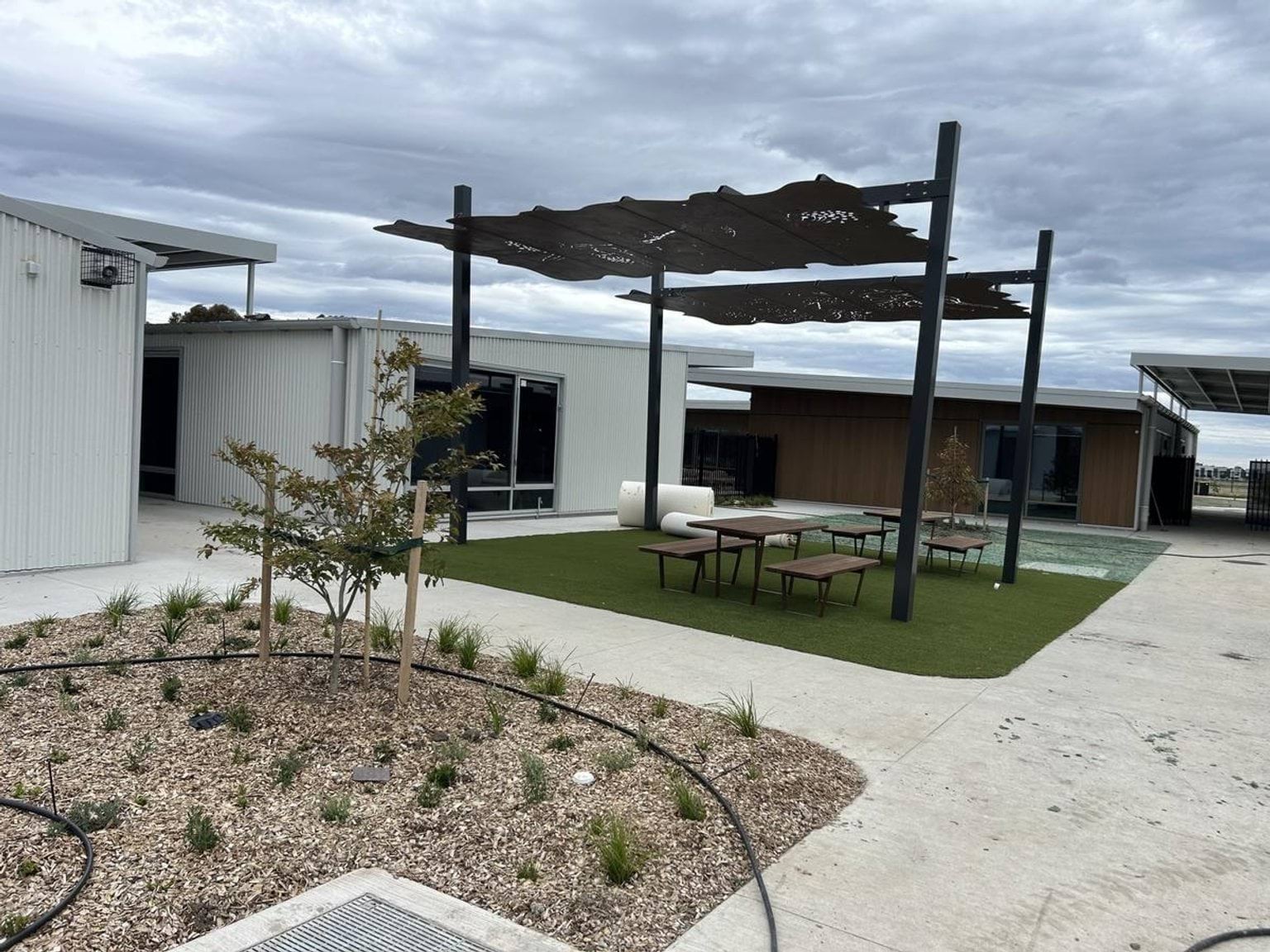 A courtyard with picnic tables on artificial grass under a decorative metal shade structure, surrounded by landscaped garden beds.