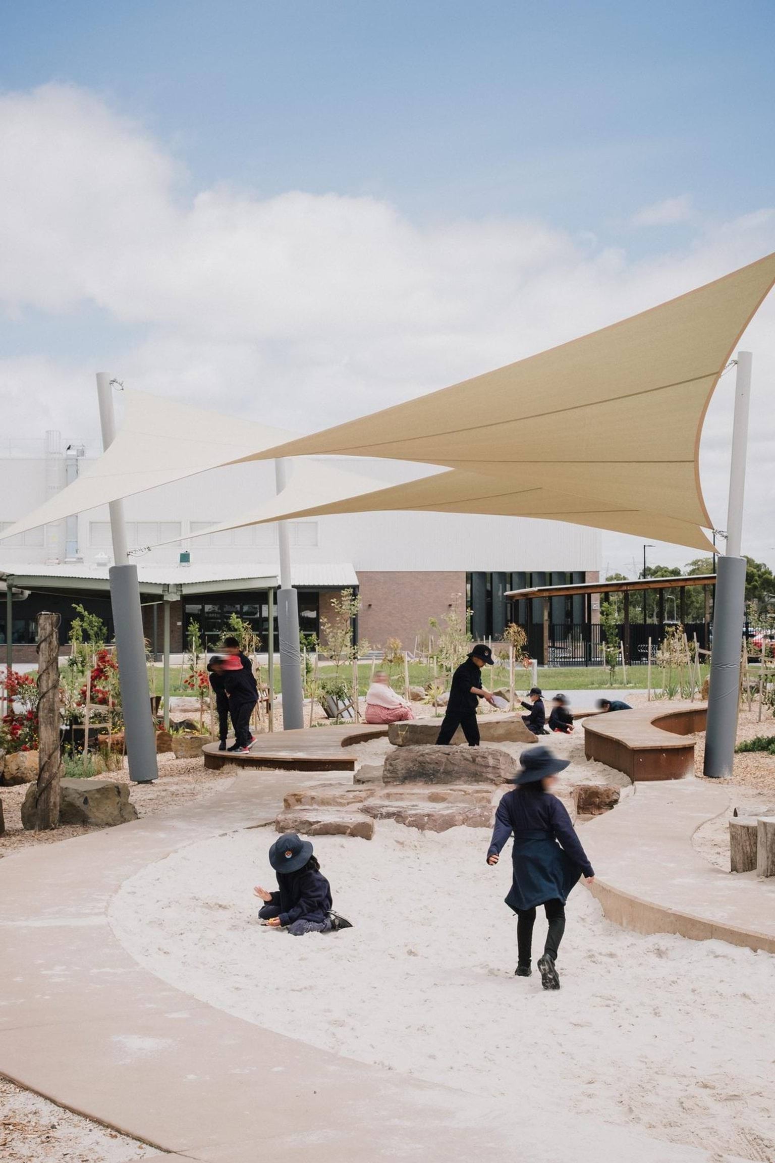 Outdoor sand play area with shade sails, natural logs, and students playing in the sand under a bright sky