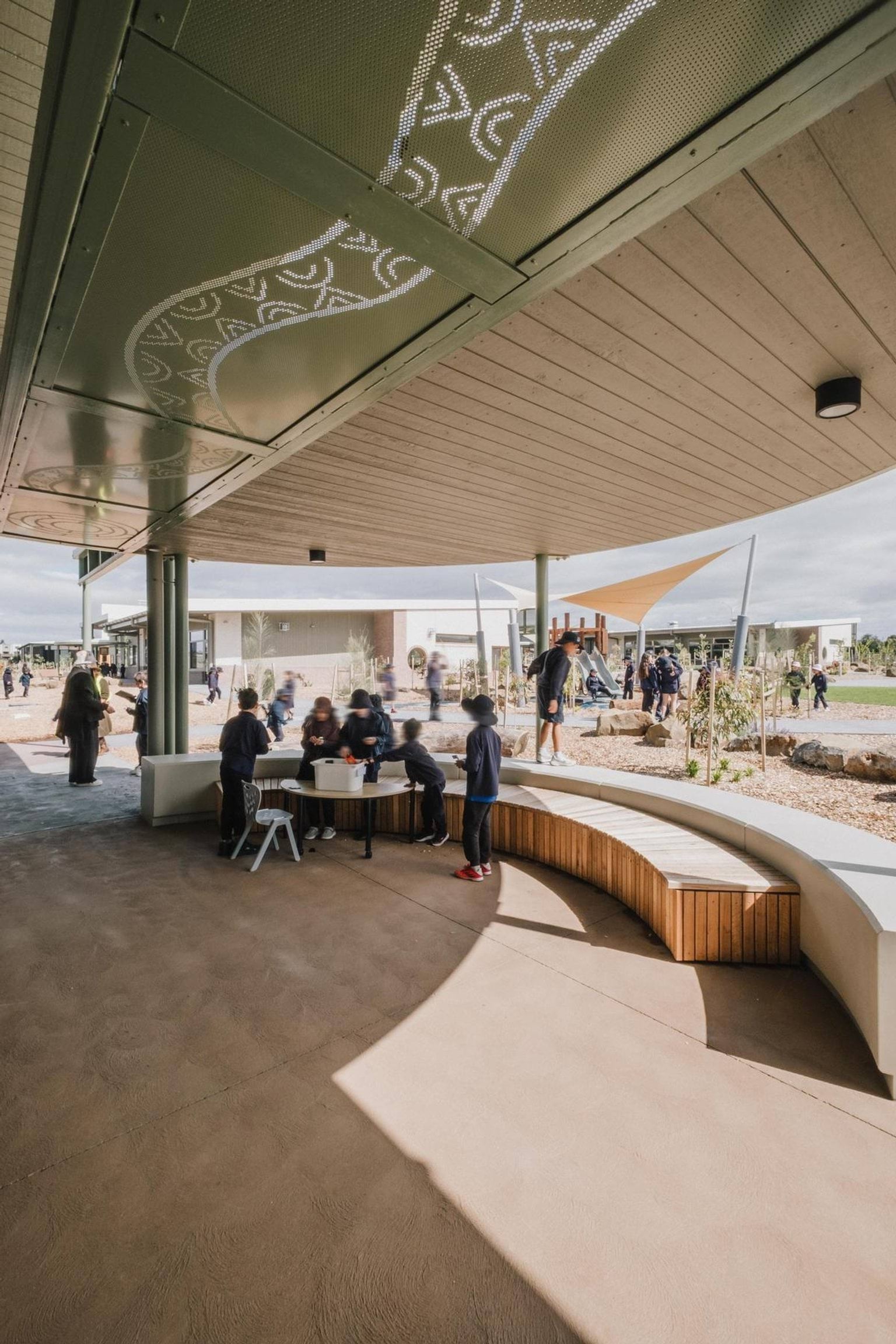 Covered outdoor learning area with curved seating, patterned ceiling panels and students gathered at a table