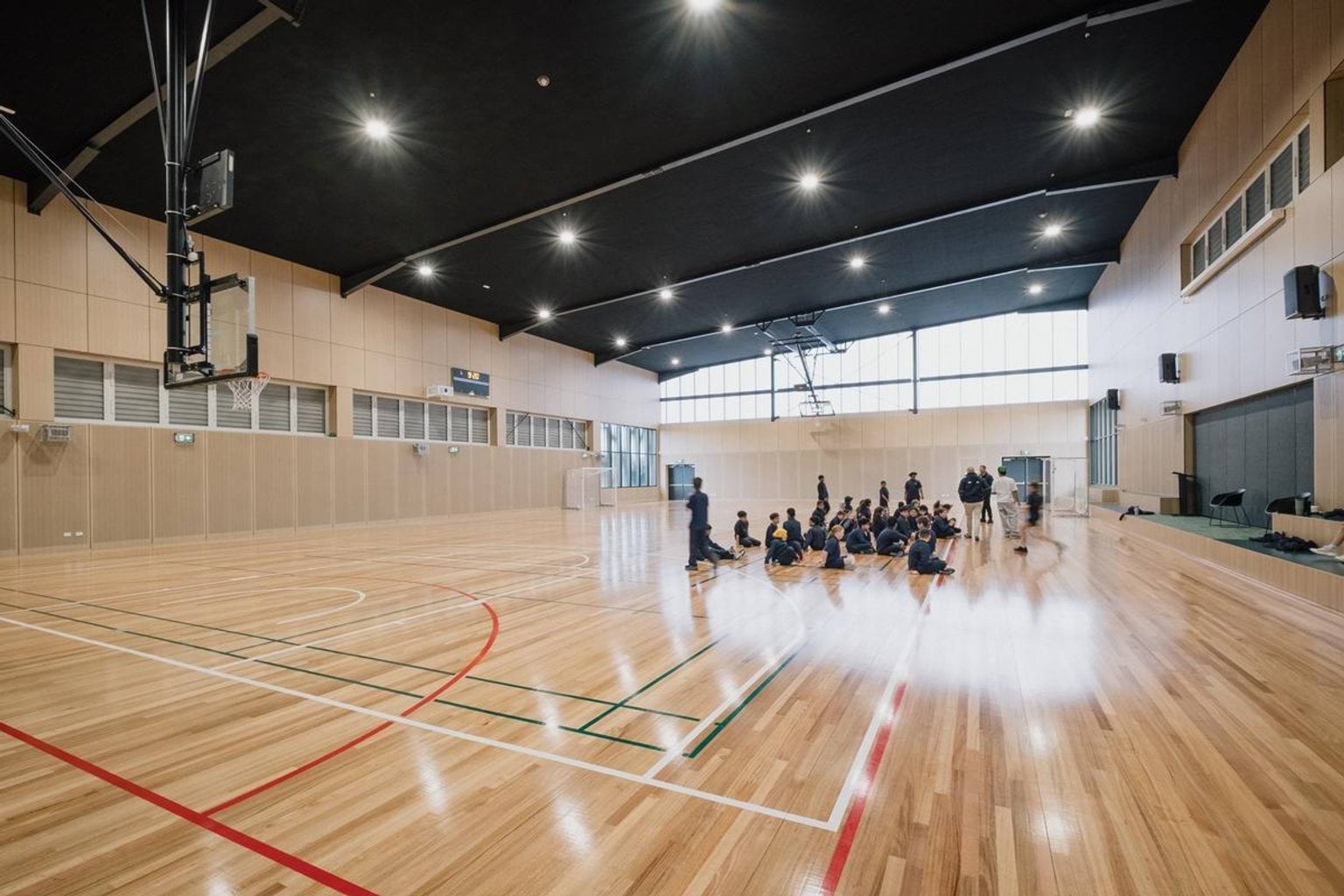 Indoor gymnasium with timber flooring, basketball hoops and students seated in a group on the court