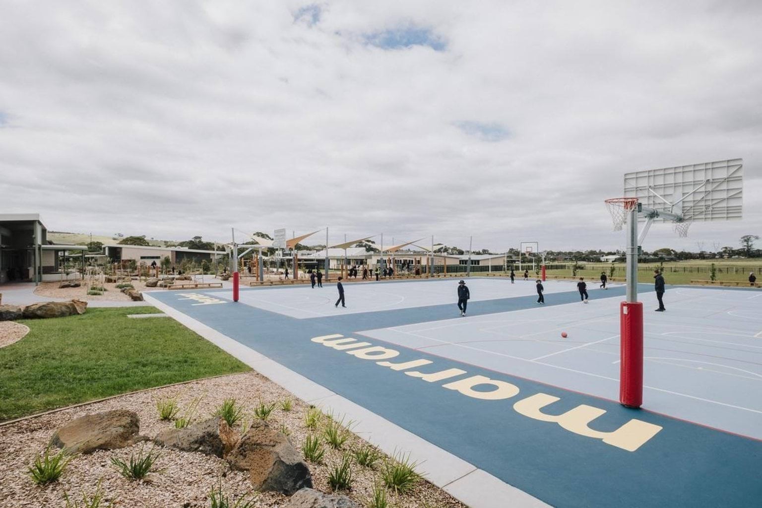 Outdoor basketball court with painted markings and hoops, with students playing in the background