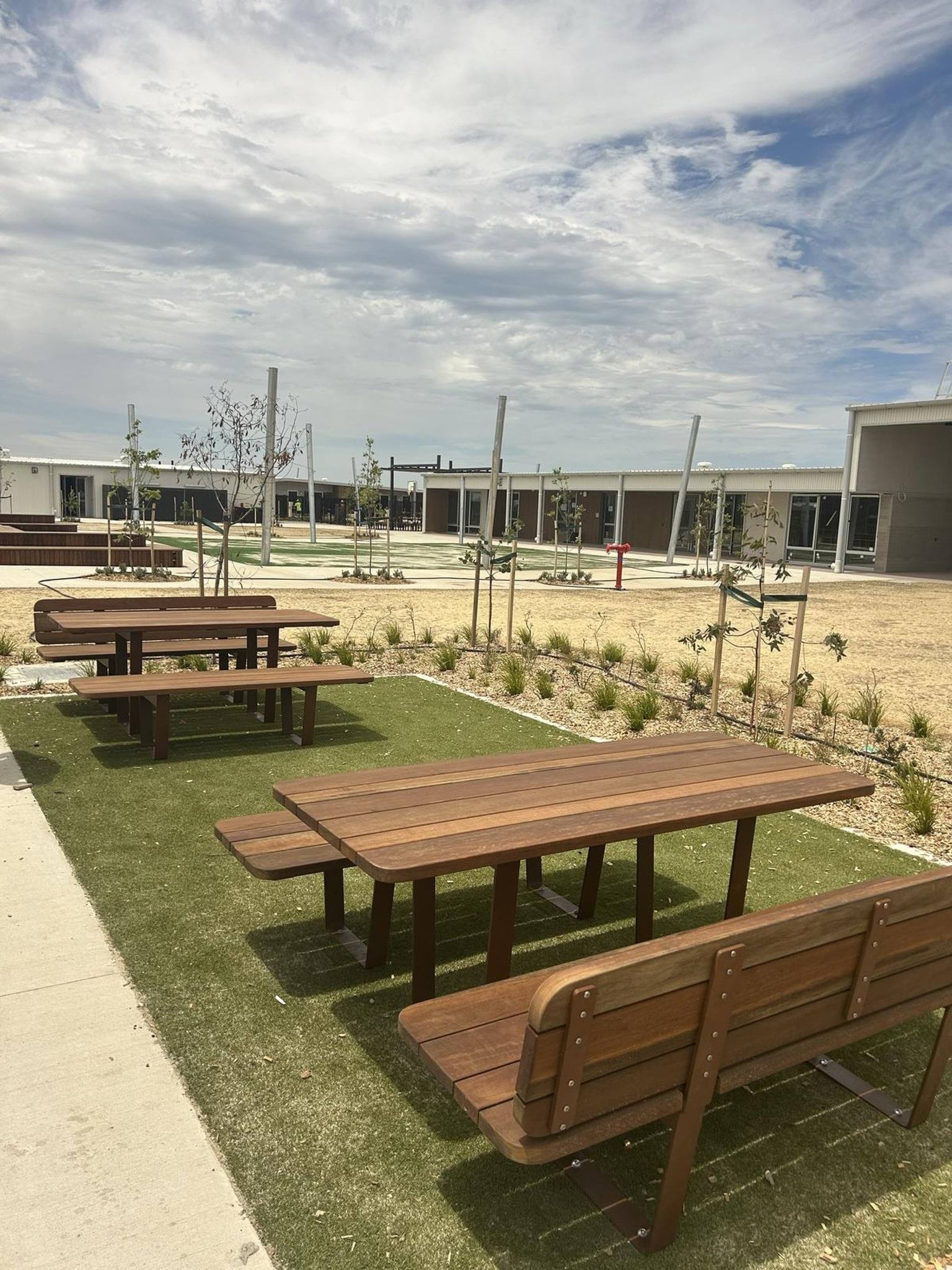An outdoor area with wooden picnic tables on artificial grass, surrounded by young trees and landscaped garden beds.