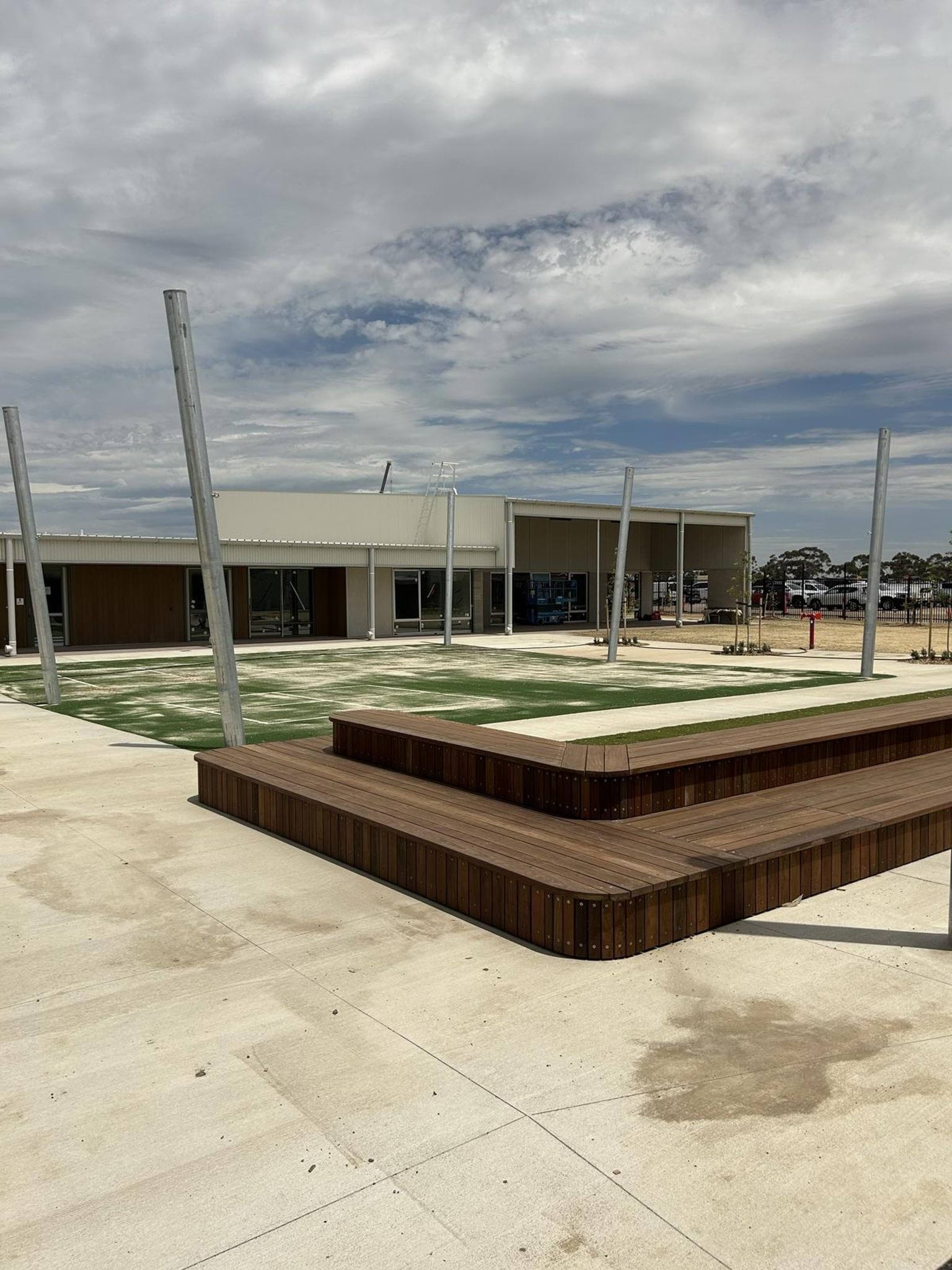 A paved courtyard with timber seating platforms and tall metal poles, in front of a modern school building with wide glass doors.