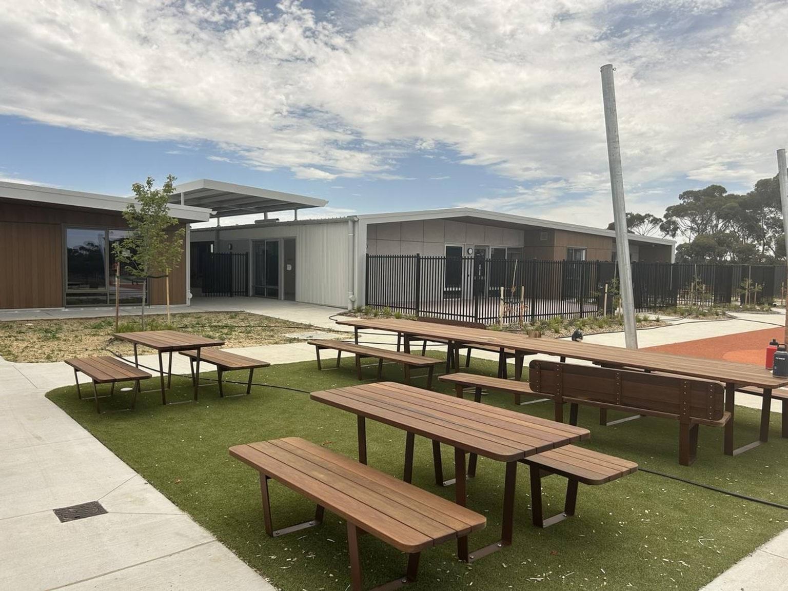 Wooden picnic tables arranged on artificial grass in a courtyard between modern school buildings with timber and grey cladding.