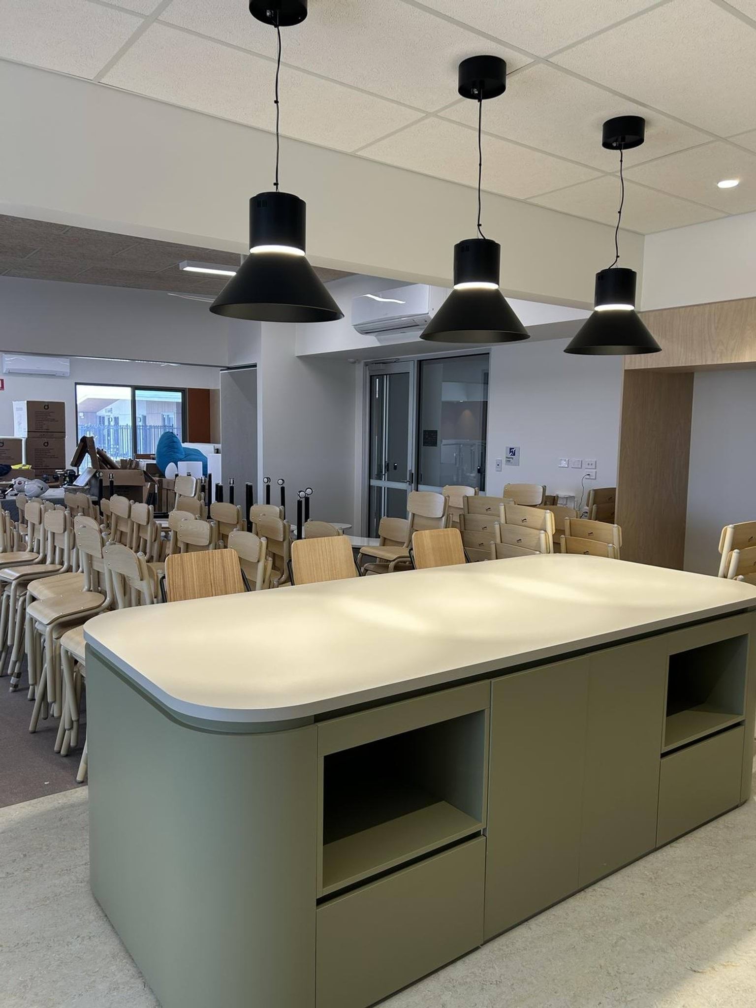 An indoor learning space with a large central island bench, rows of stacked chairs, and three black pendant lights hanging from the ceiling.