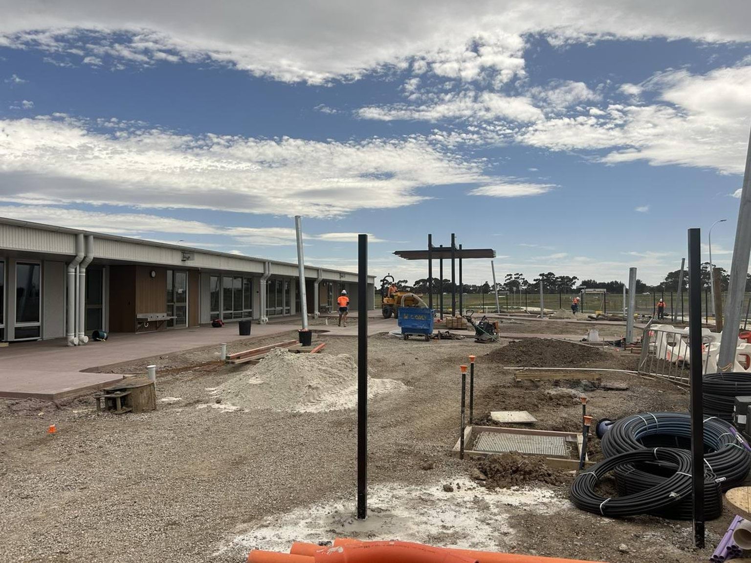 A kindergarten building with a covered walkway and large windows. The foreground shows construction work with exposed soil, pipes, and metal posts under a partly cloudy sky.