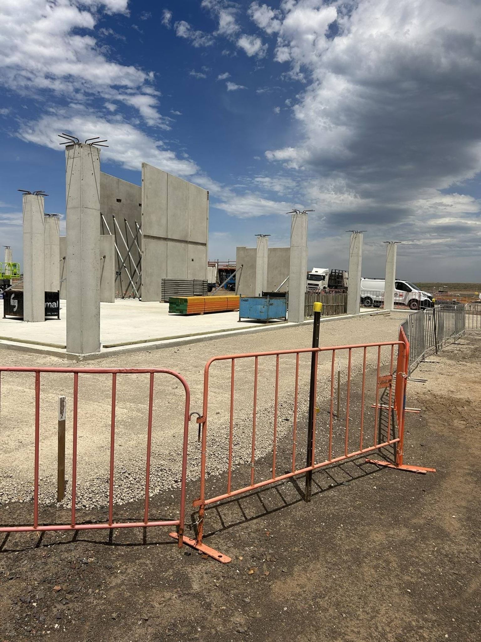 Concrete wall panels and columns forming the outline of a building, surrounded by orange safety fencing and construction materials.