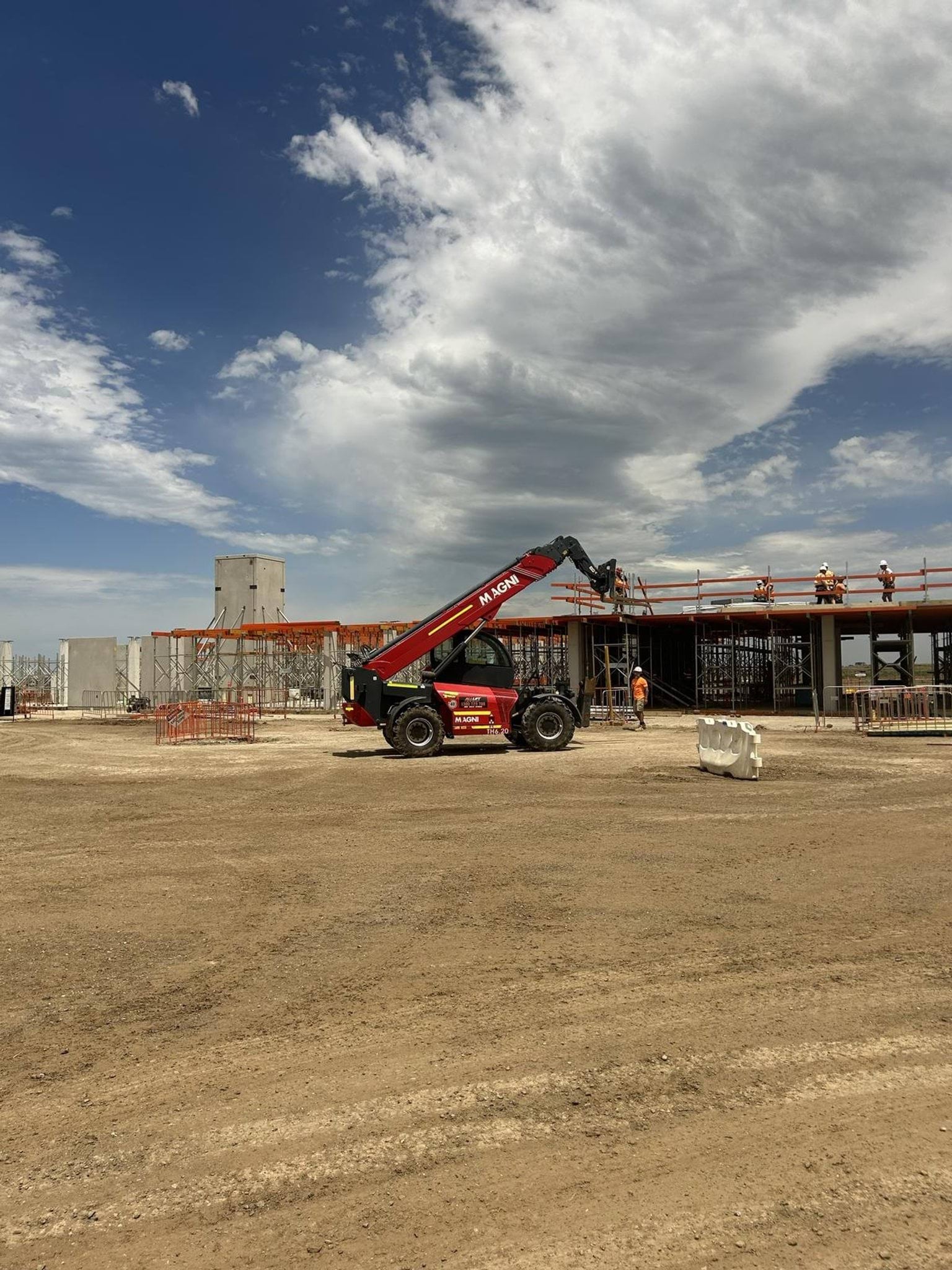 A construction site with a partially built school structure, workers on scaffolding, and a red telescopic handler in front under a partly cloudy sky.
