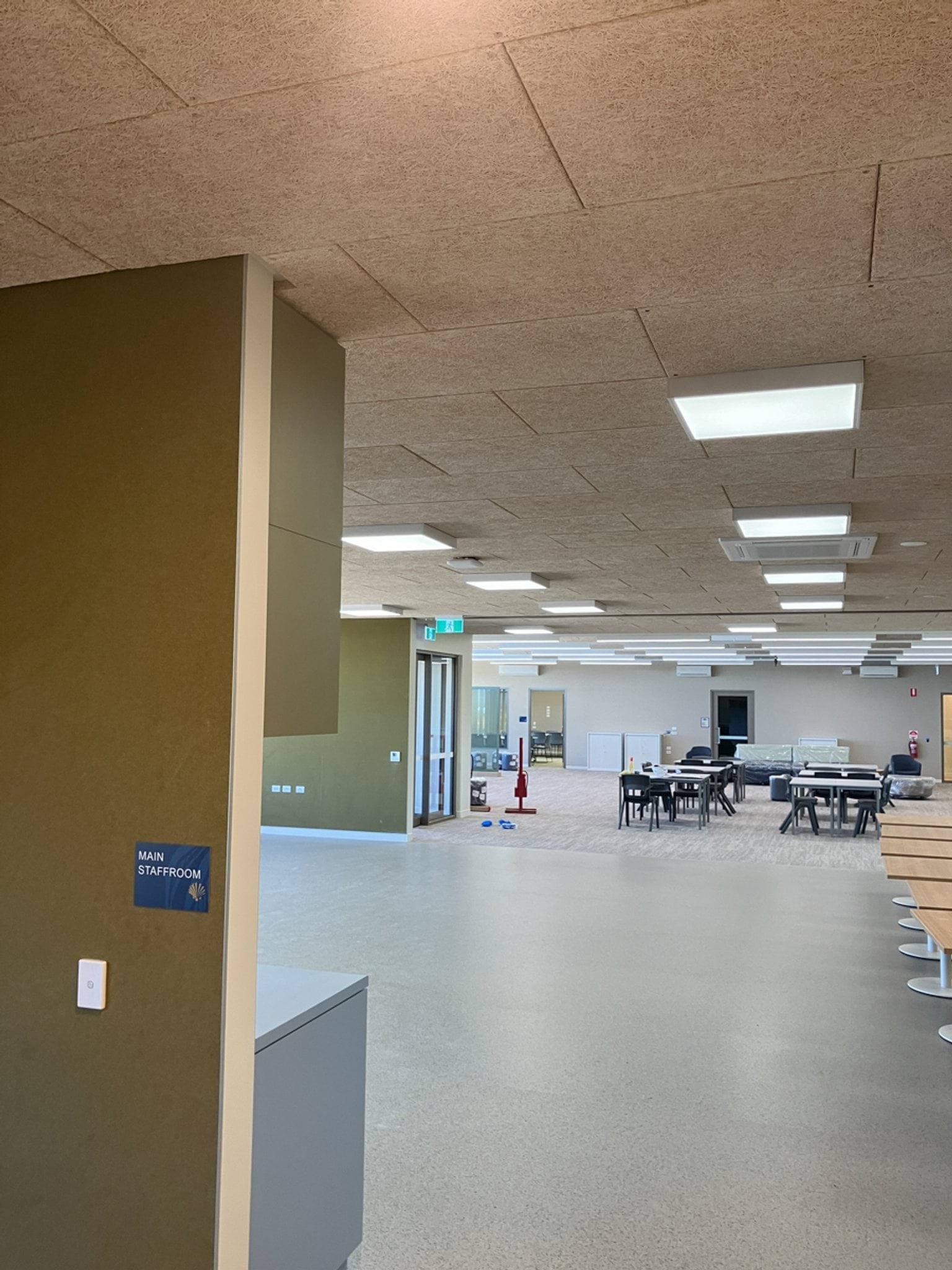 An unfinished staffroom with exposed flooring, tables arranged in the centre, and ceiling panels installed. Construction tools are visible.
