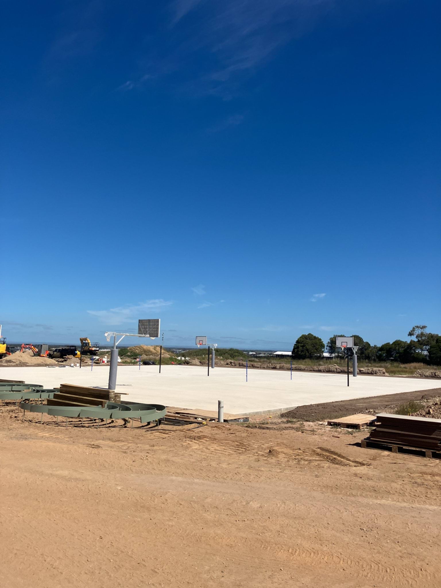 Two outdoor basketball courts with concrete slabs and hoop posts installed, surrounded by dirt and construction equipment.