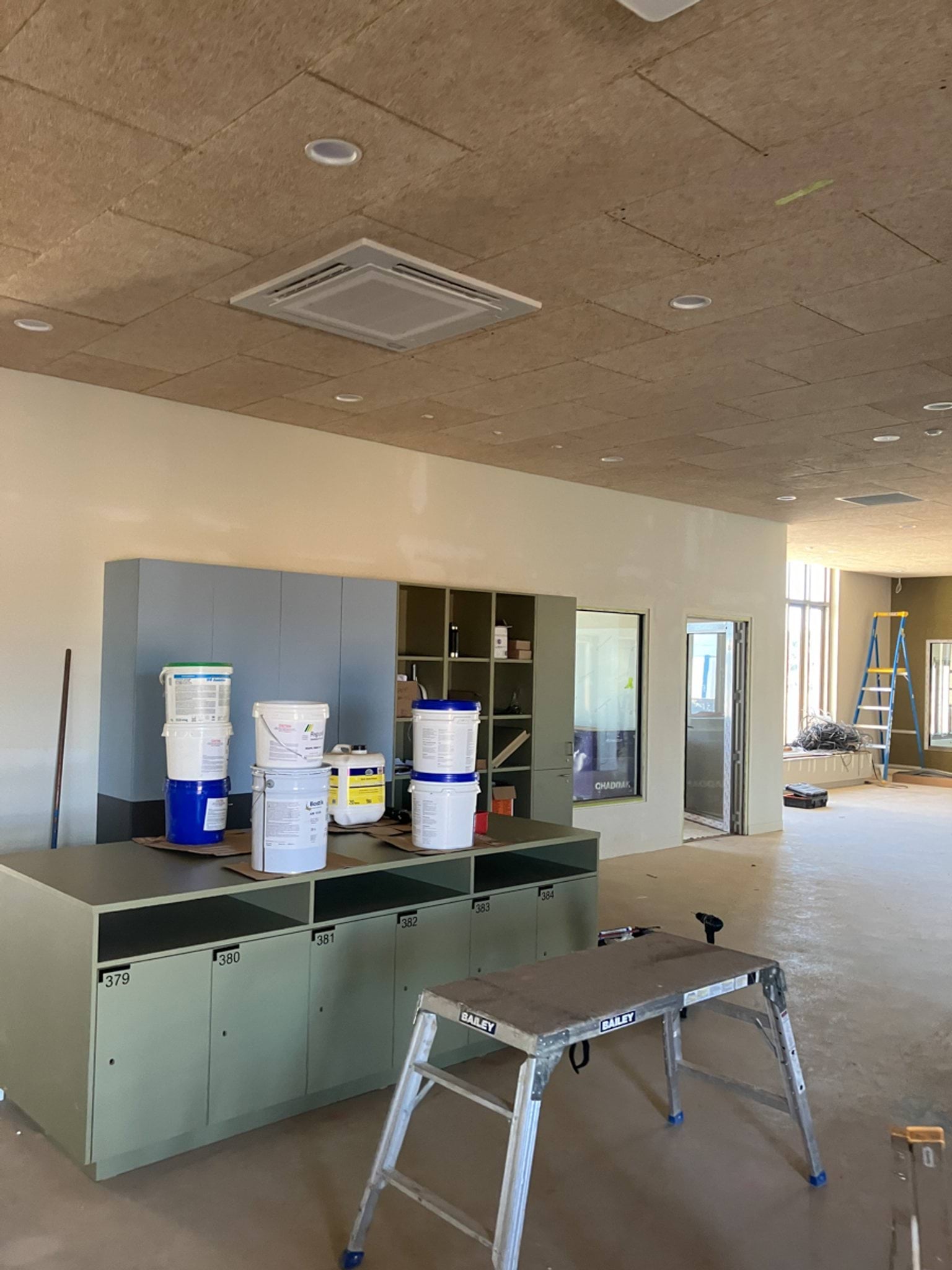 A partially completed storage area with cabinetry, paint buckets, and a step ladder. Large windows provide outdoor views.