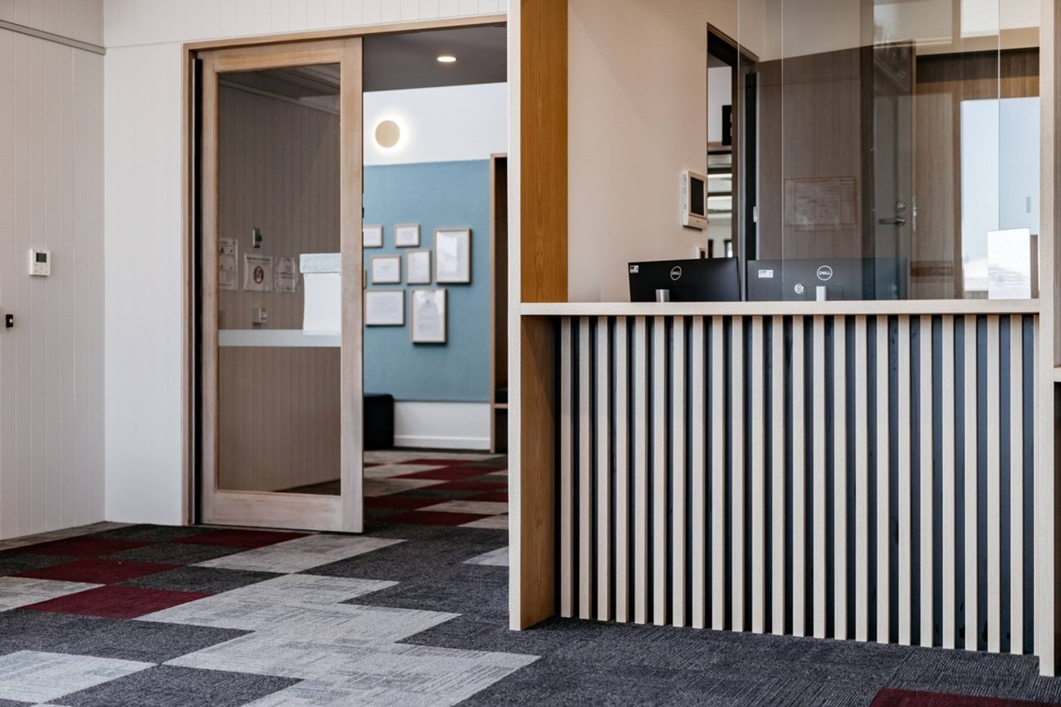 A school reception area with a timber-framed glass door, a counter featuring vertical timber slats, and patterned carpet in grey, white, and burgundy tones. Certificates are displayed on a blue wall in the background.