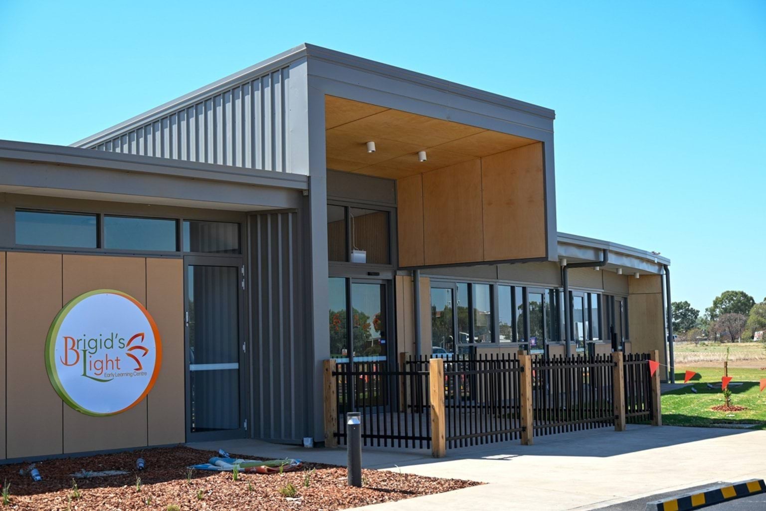 The exterior of a modern school building with grey and timber panels, large windows, and a covered entry. A round sign reads “Brigid’s Light Early Learning Centre,” and the area is landscaped with mulch and plants.