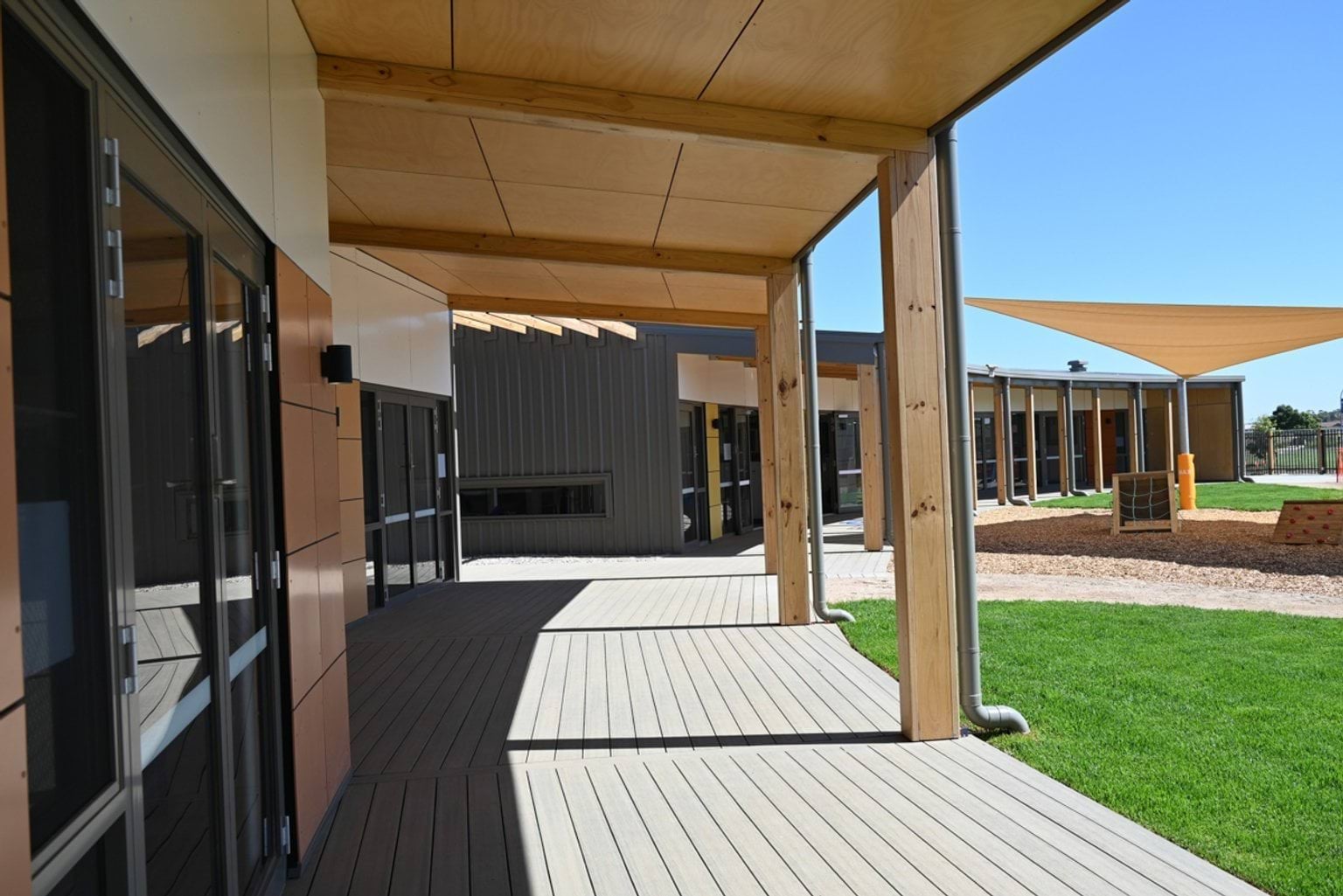A covered outdoor walkway with timber posts and decking, connecting classrooms. The area overlooks a green lawn and a playground with shade sails.