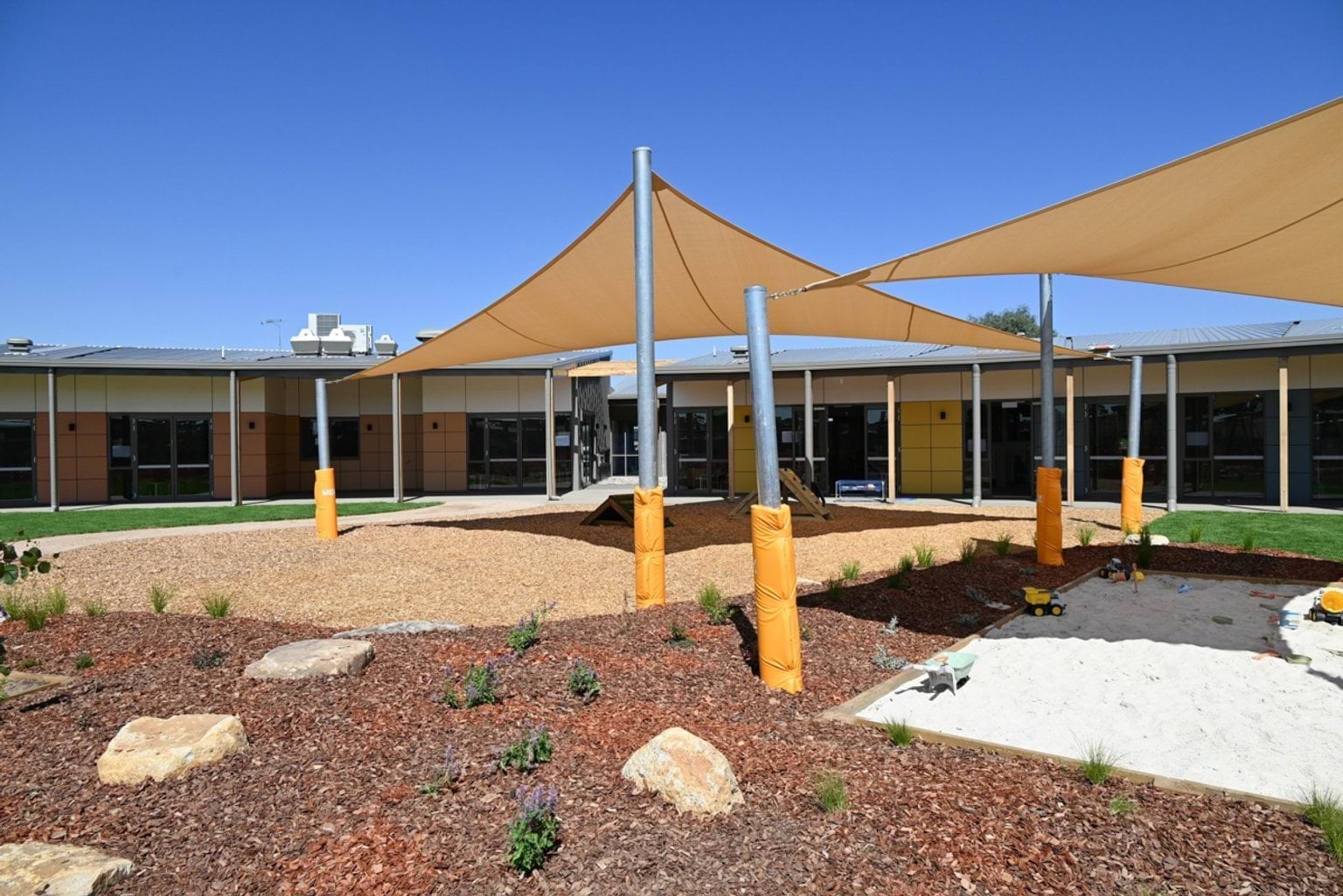 An outdoor play area with tan shade sails, a sandpit, and landscaped garden beds with rocks and plants. The surrounding building has large windows and a mix of cream and brown panels.