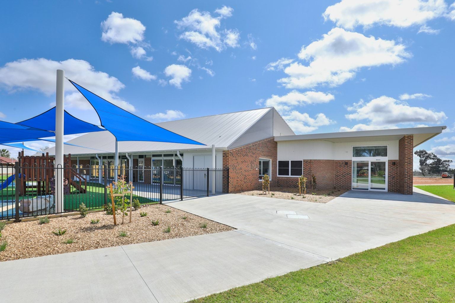 The exterior of a school building with brick and white panel walls, large windows, and blue shade sails covering the outdoor play area.