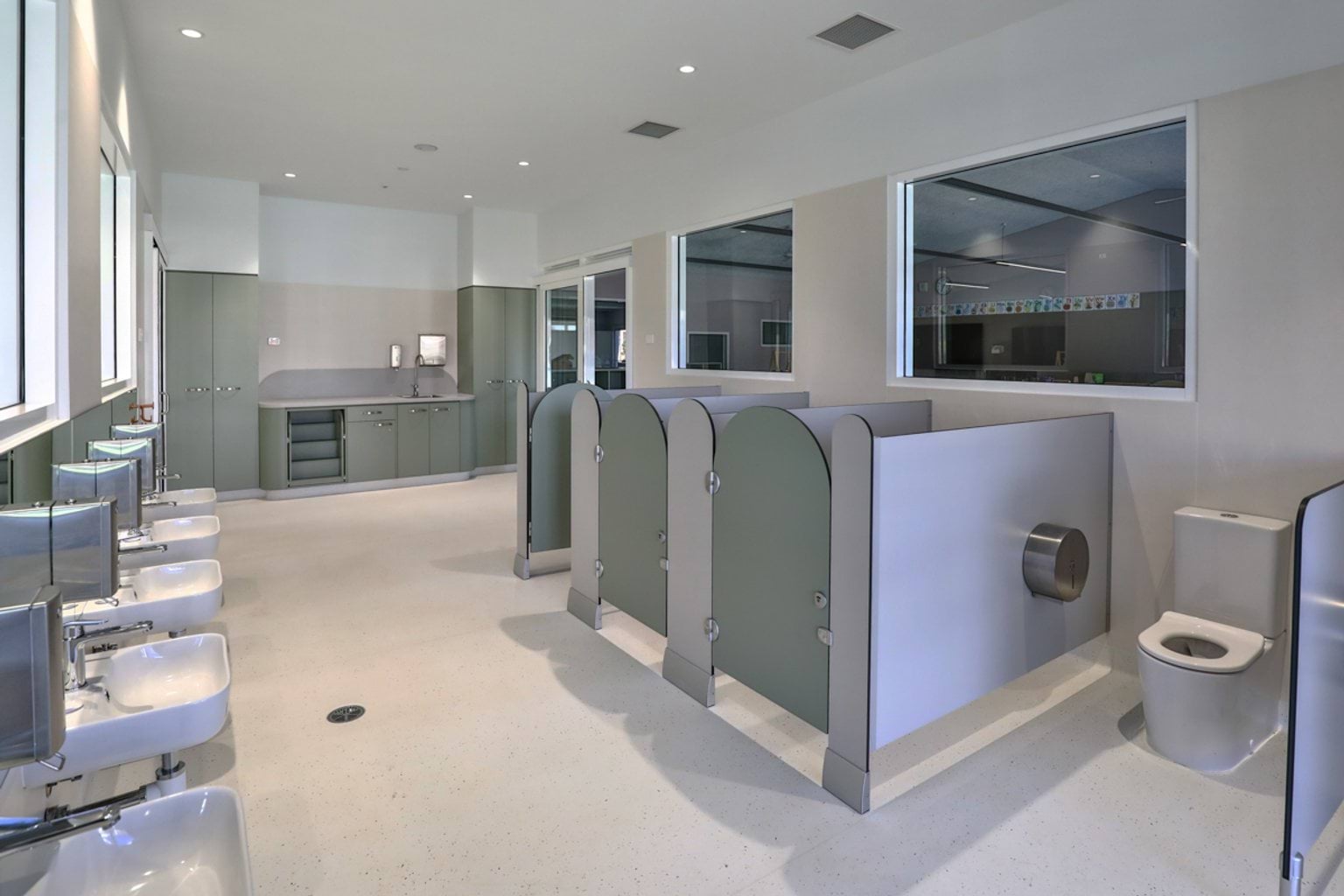 A children’s bathroom with low-height toilet cubicles, basins along one wall, and storage cabinets at the rear.