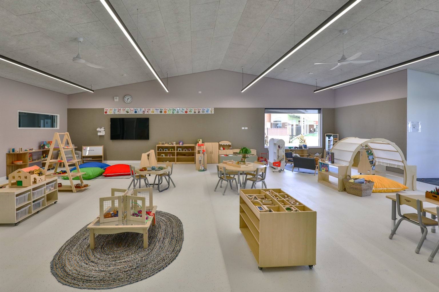 A light-filled early learning classroom with small tables, timber play furniture, and a variety of activity stations.